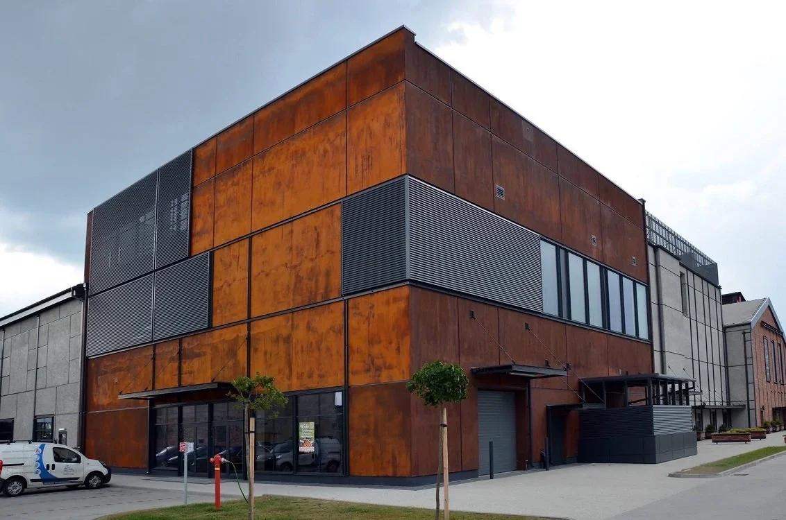 Modern multi-story building with a rust-colored metal exterior and large windows, situated on a street with parked cars and small trees.