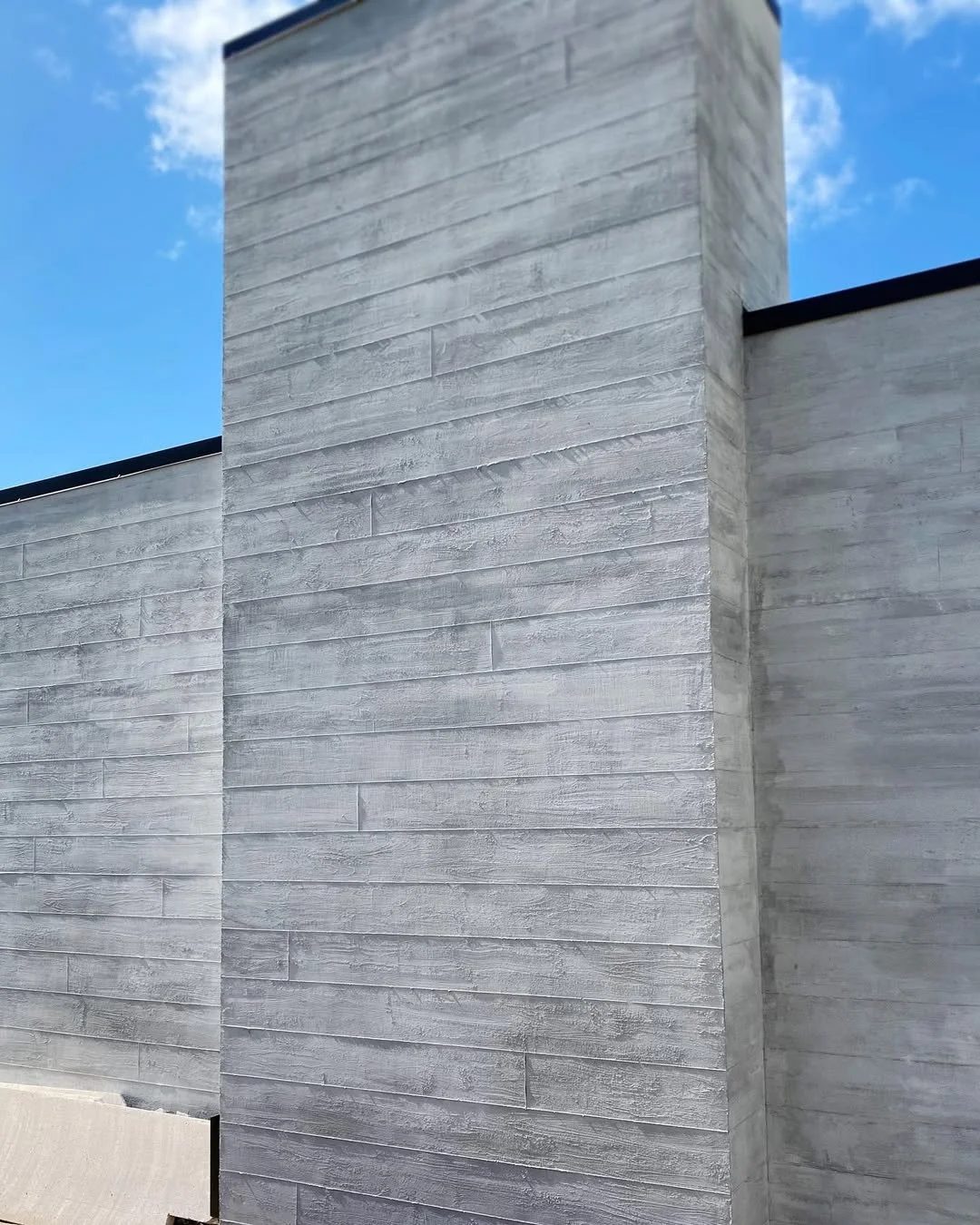 Close-up view of a modern exterior wall made of grey textured concrete panels under a blue sky with some clouds.