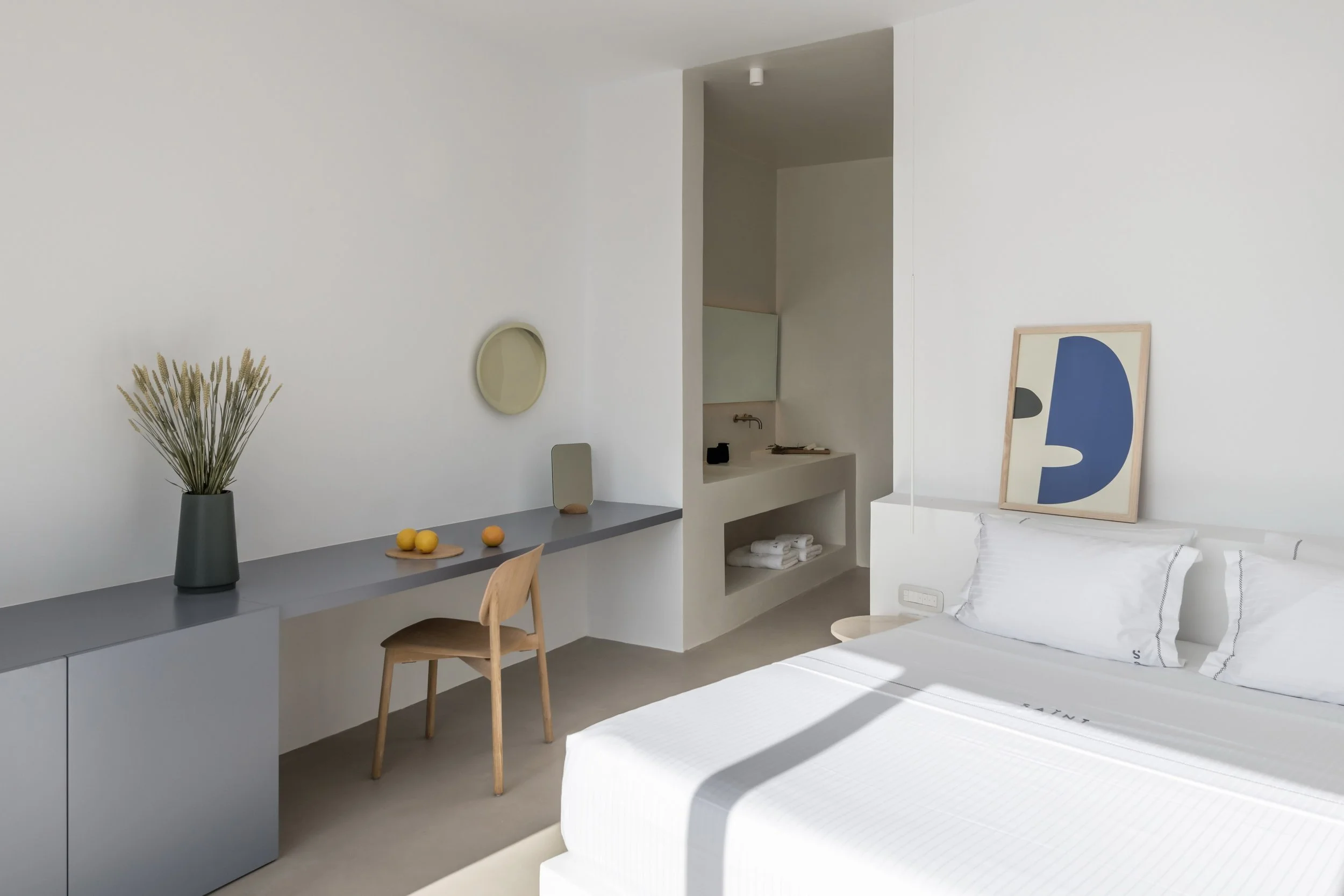 Minimalist bedroom with white bed, framed abstract art, wooden desk with vase of dried plants, small mirror, and a corner sink with towels.