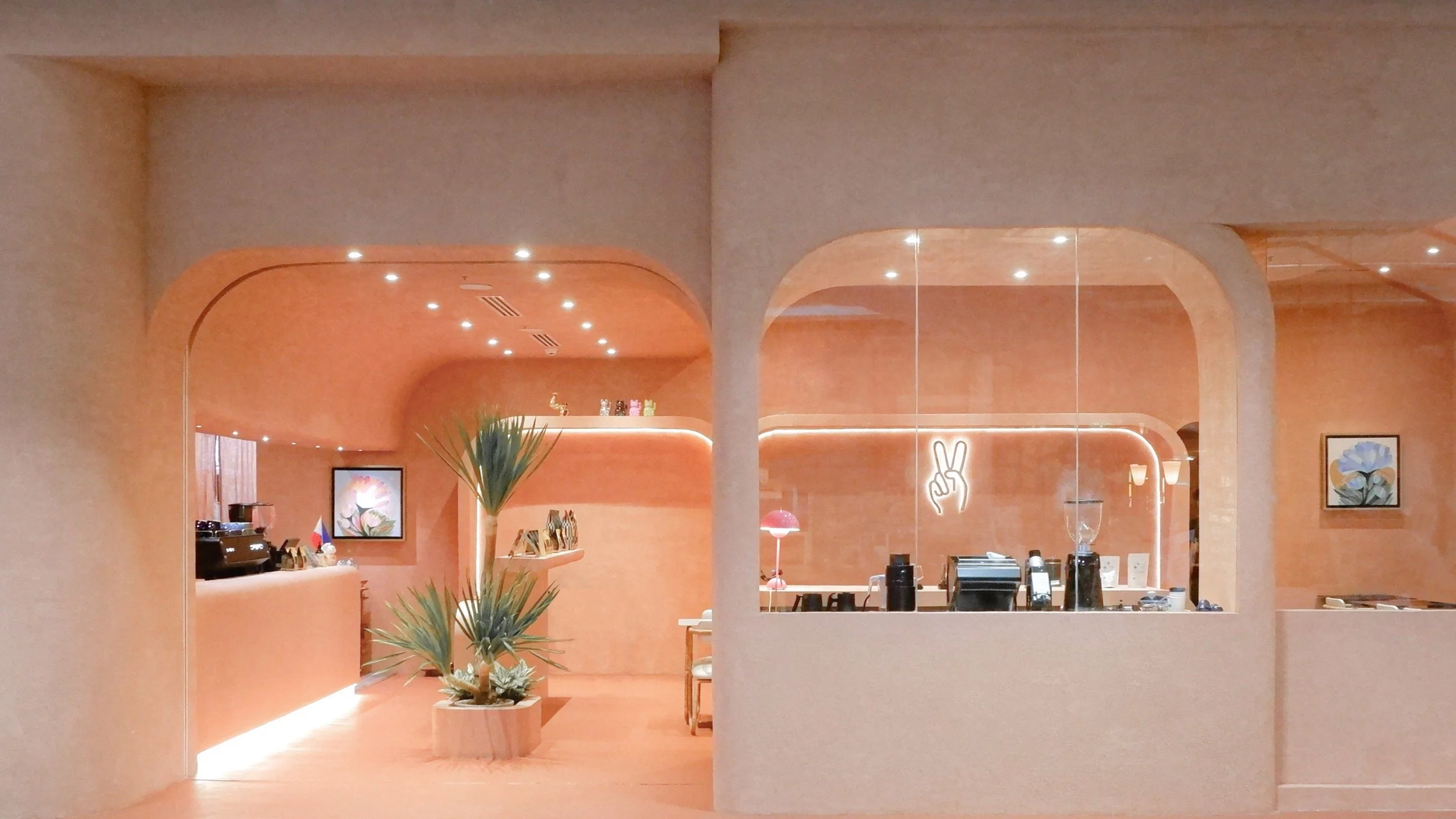 Interior of a modern, warmly lit cafe with peach-colored walls, arches, and decorative art on the walls, including a neon peace sign and paintings of flowers. There are plants and a counter with various items and a mirror reflecting part of the space.