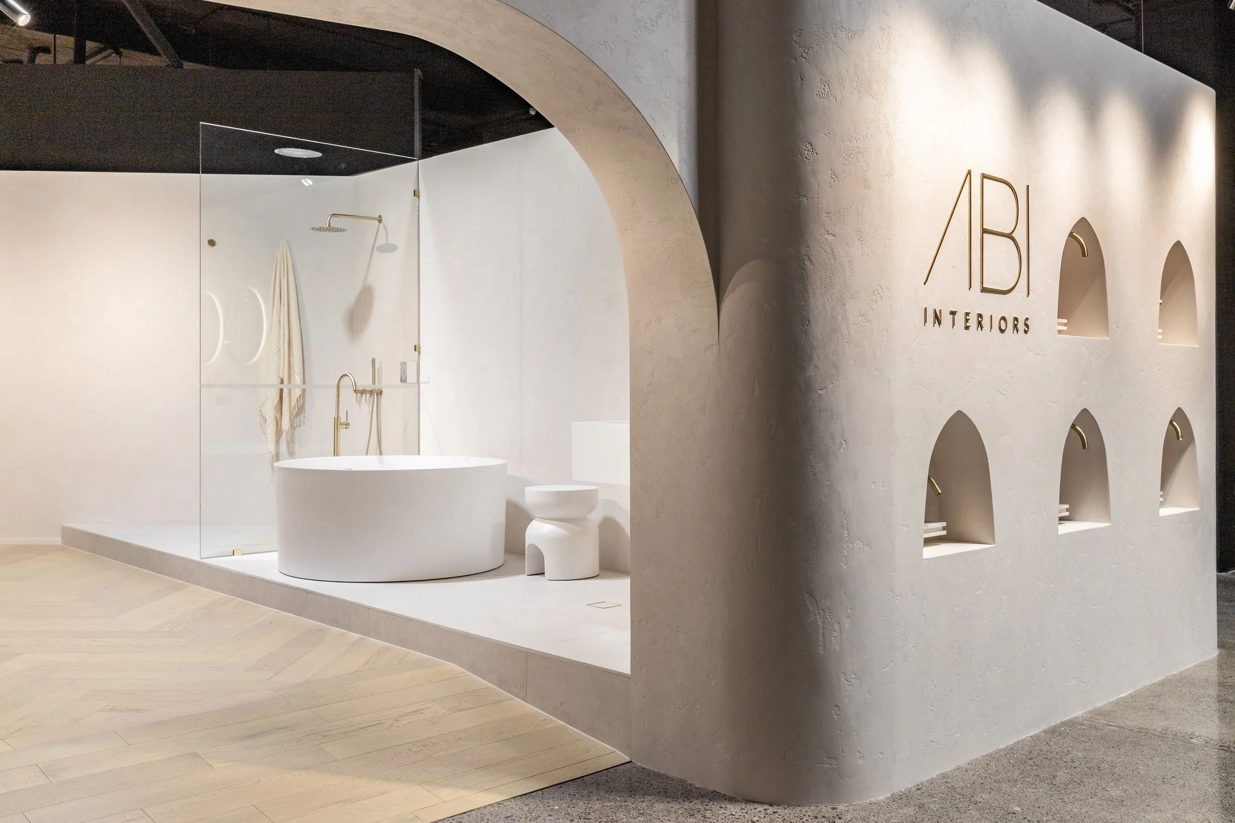 Display of a bathroom interior with a white freestanding bathtub, a towel hanging on the wall, a butler sink, and a small stool, surrounded by a glass partition.