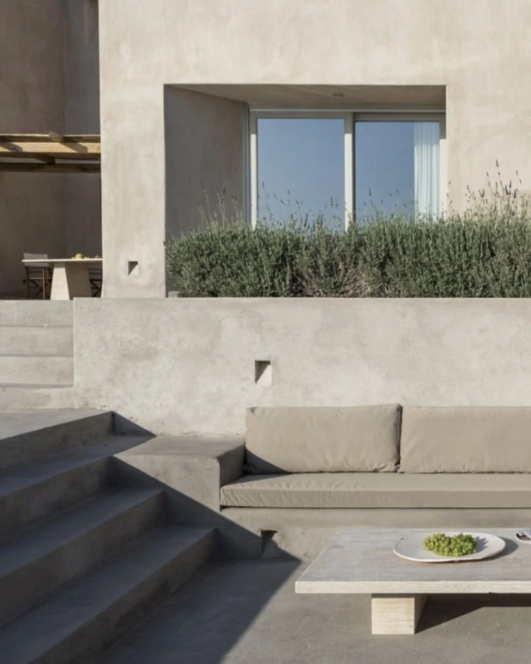 Modern outdoor patio with built-in concrete sofa, steps, and a low table with a bowl of grapes, against a beige stucco wall with a large window and green shrubbery.