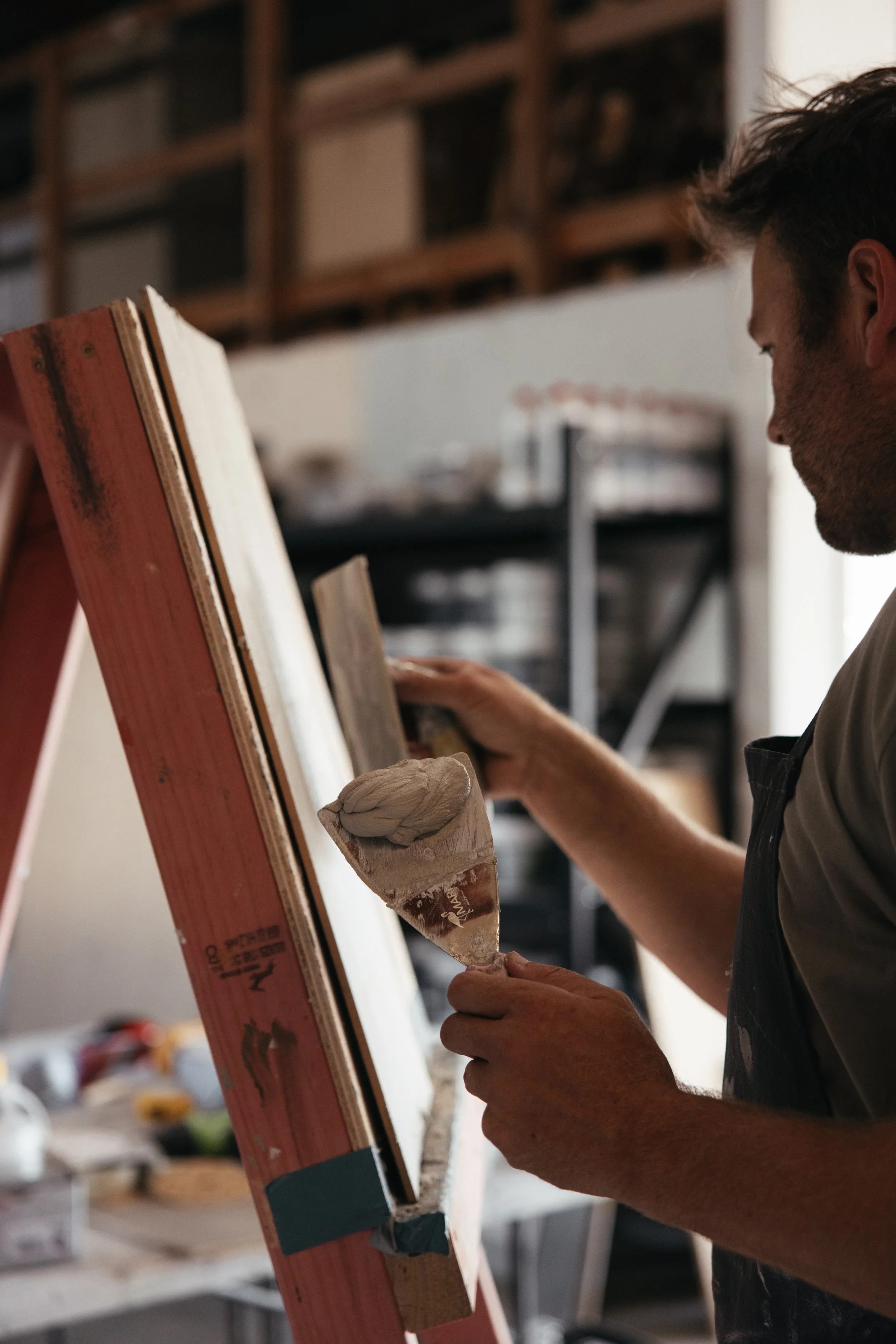 A man with dark hair is working on a piece of wood at an art studio or workshop. He is holding a putty knife with some white material on it, and appears to be preparing or shaping the wood.