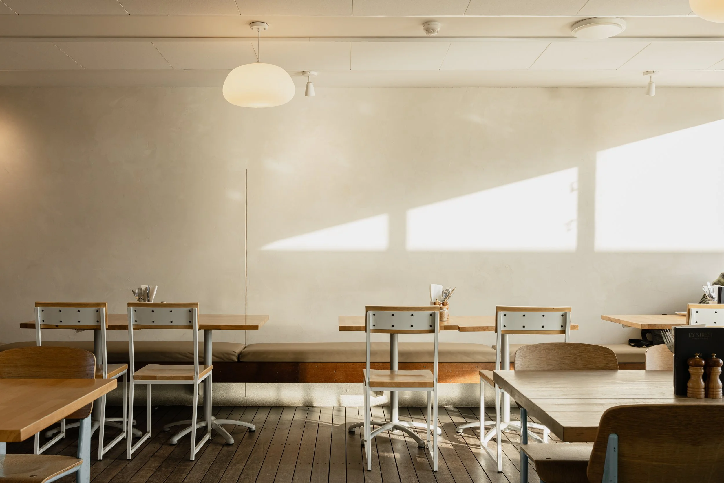 Empty dining area in a restaurant with tables, chairs, and sunlight coming through windows, creating shadows on the wall.