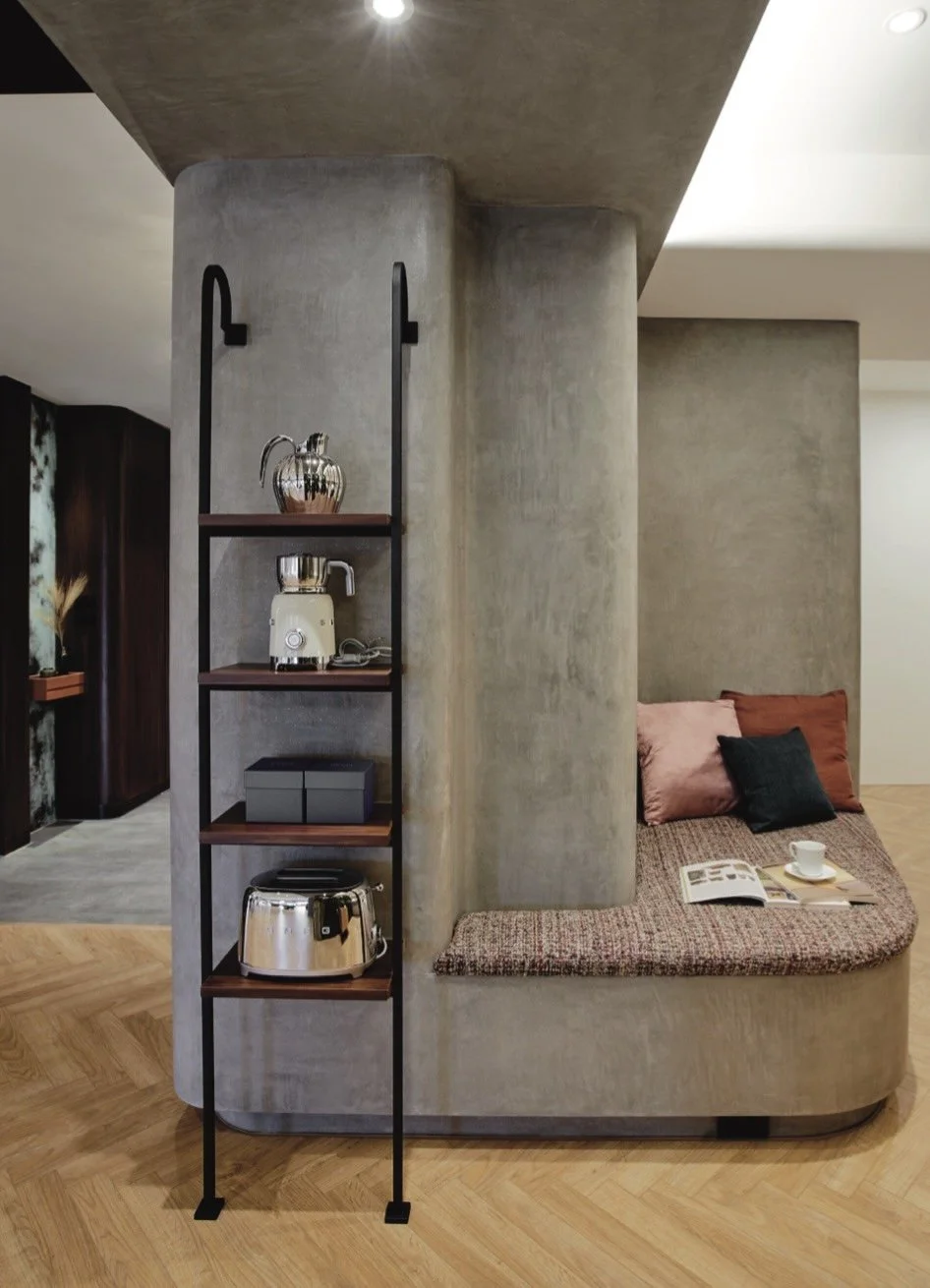 Small shelving unit with a teapot, toaster, and decorative boxes next to a curved, built-in seating area with pillows, a book, and a cup in a modern interior.