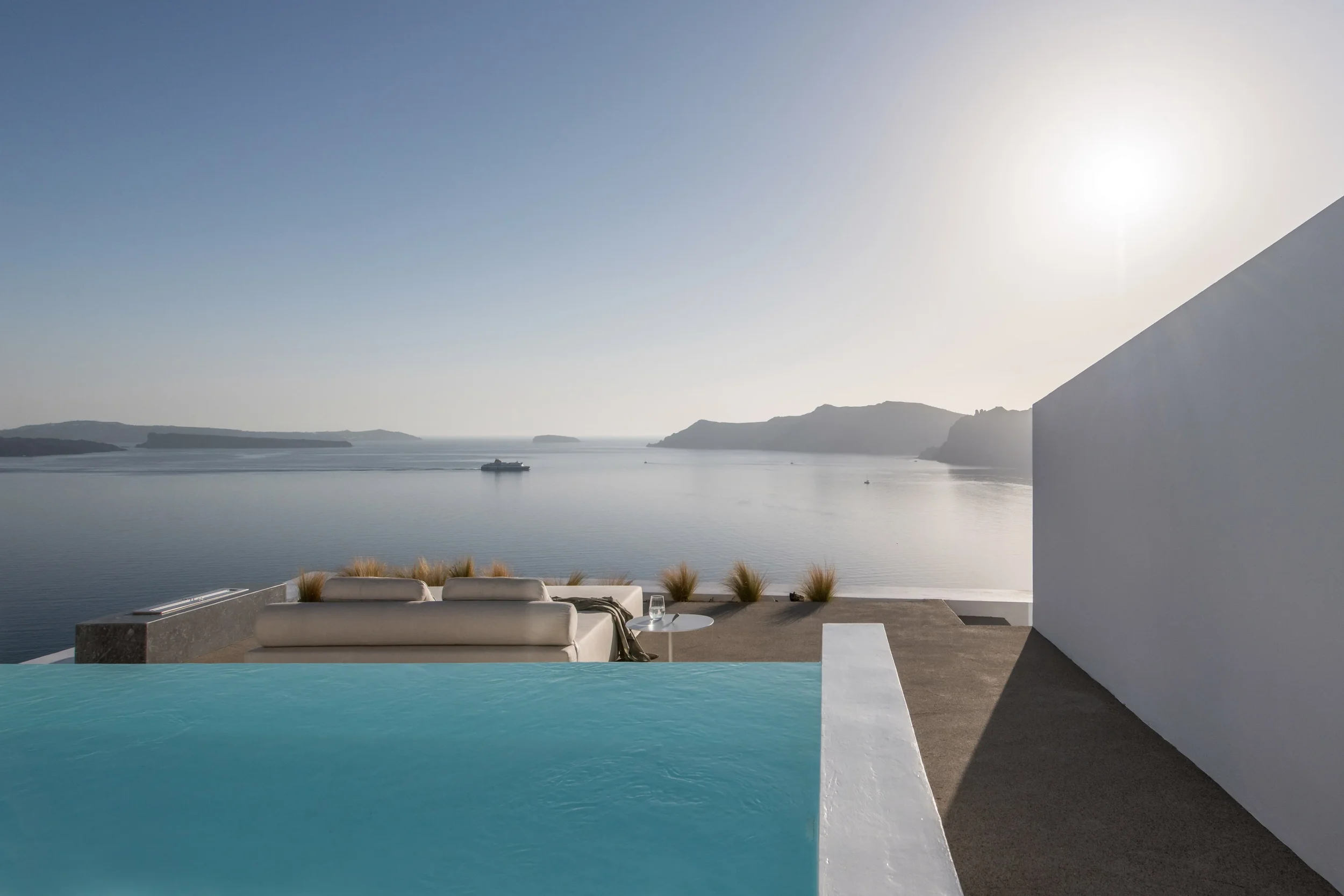 A scenic view of a large body of water with distant islands and mountains, seen from a modern outdoor pool area with white outdoor furniture and decorative plants, against a clear blue sky and bright sunlight.