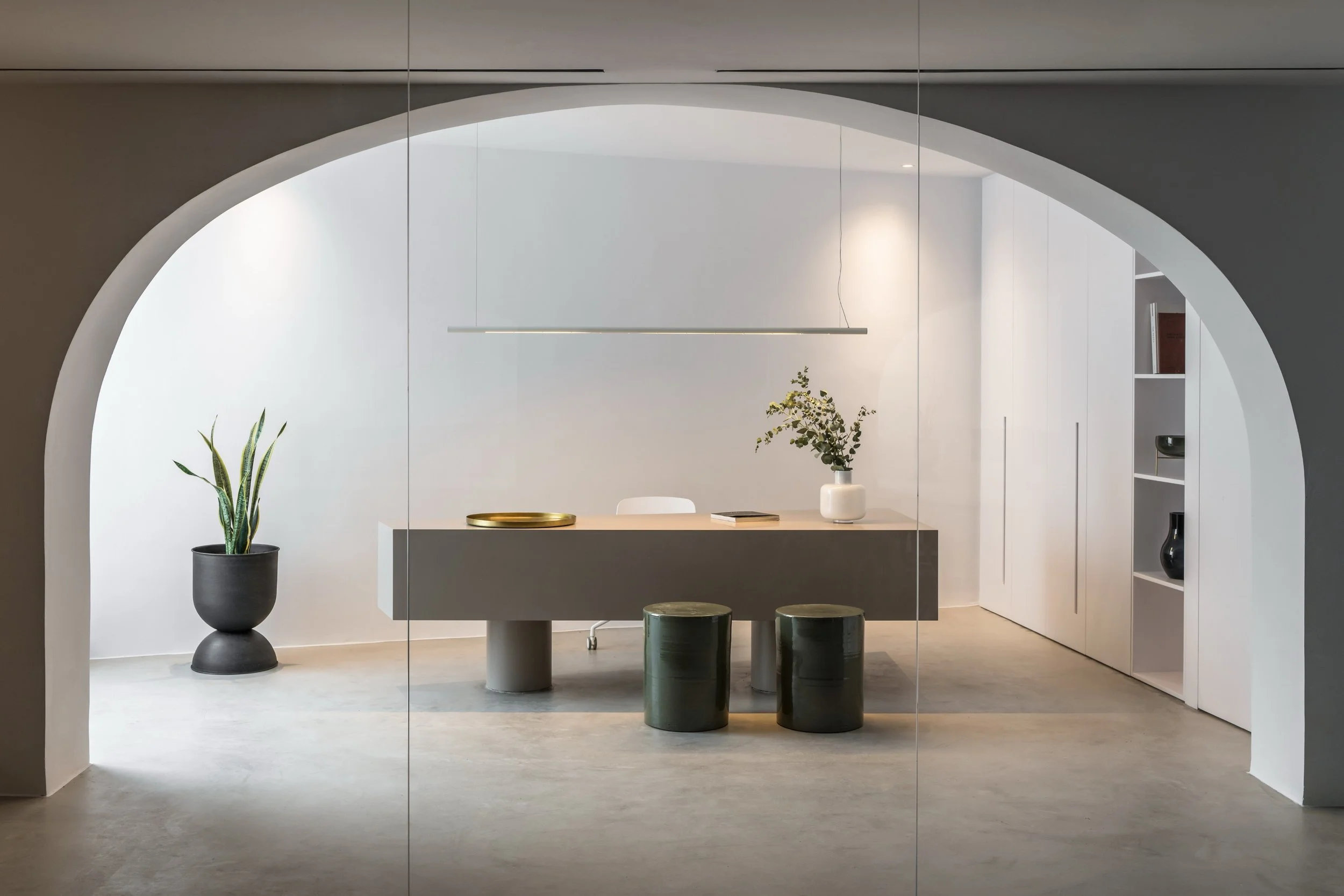 Minimalist interior space featuring a white and grey dining or work table with green and white stools, a potted plant, a white vase with greenery, a gold tray, a book, and a sleek hanging light fixture, framed by an arched entryway.