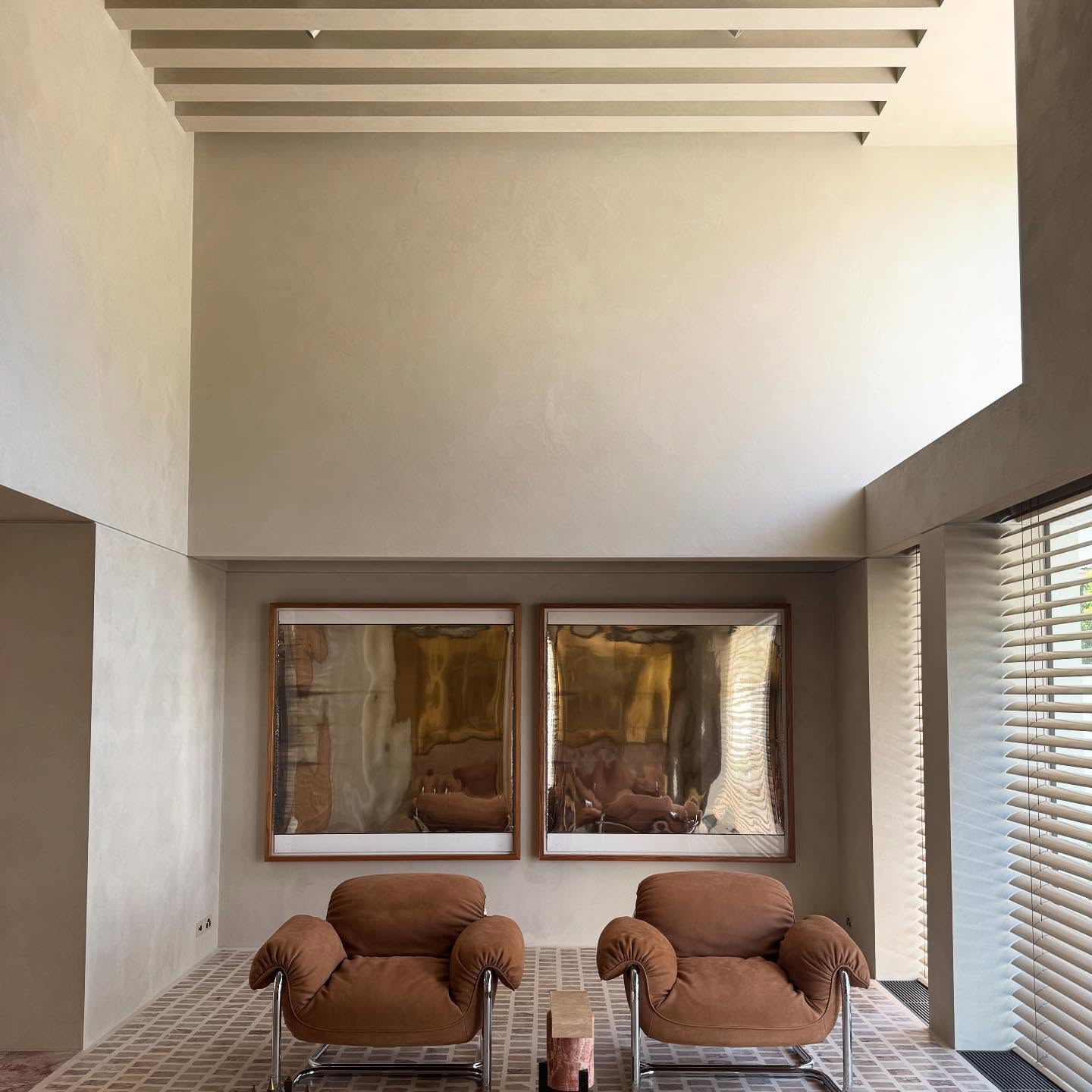 Living room area with two brown cushioned armchairs, a small wooden table, wall art with two large reflective framed paintings, beige walls, and window blinds.