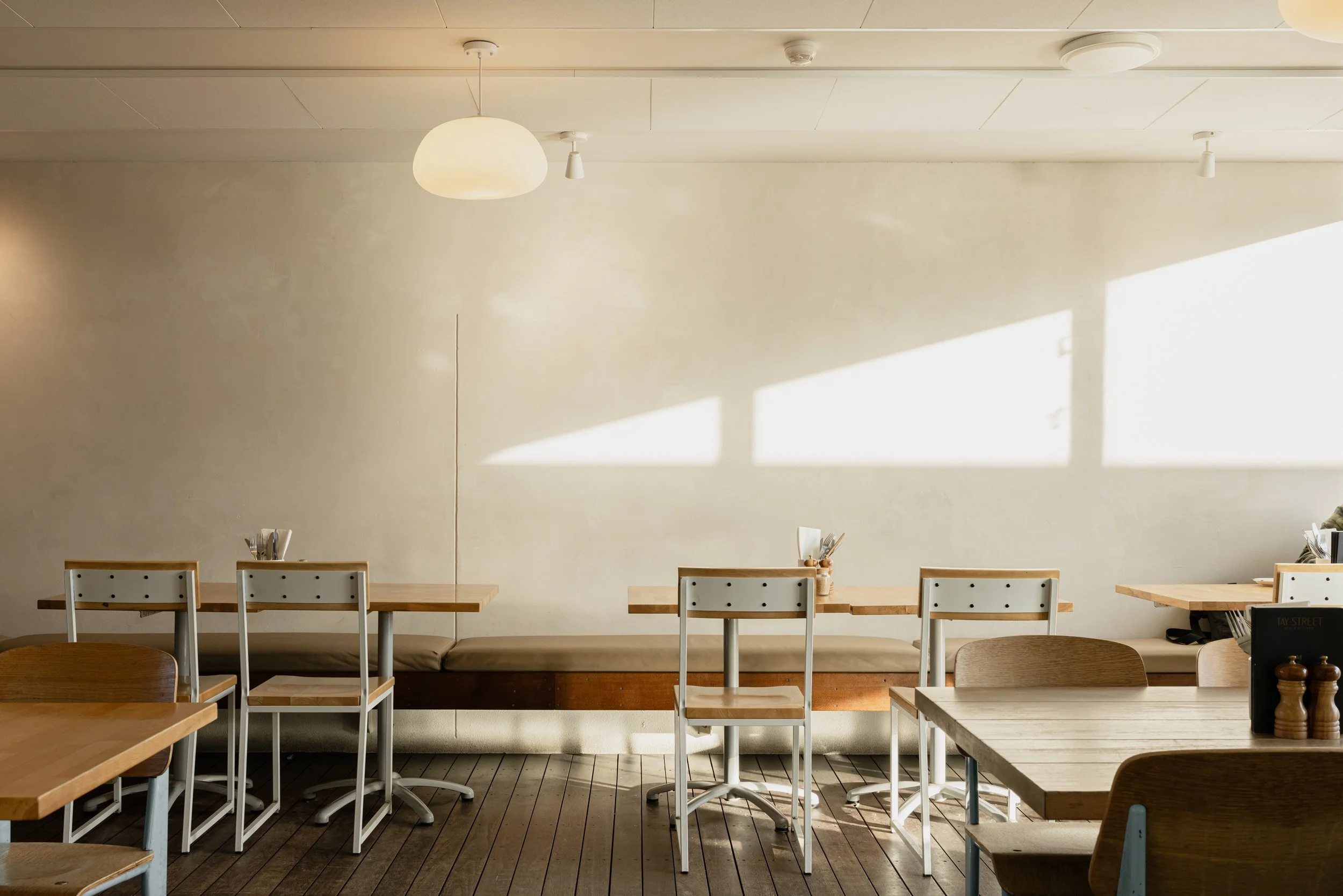 Interior of a restaurant with wooden tables and white chairs, sunlight casting shadows on a plain beige wall, ceiling lights, and table settings.