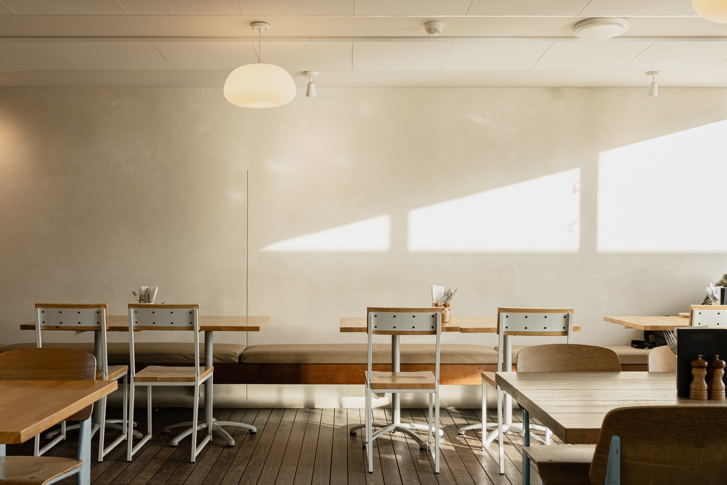 Interior of a dining area with a long table, wooden chairs with white backs, and a simple neutral wall. Sunlight and shadows create a geometric pattern on the wall.