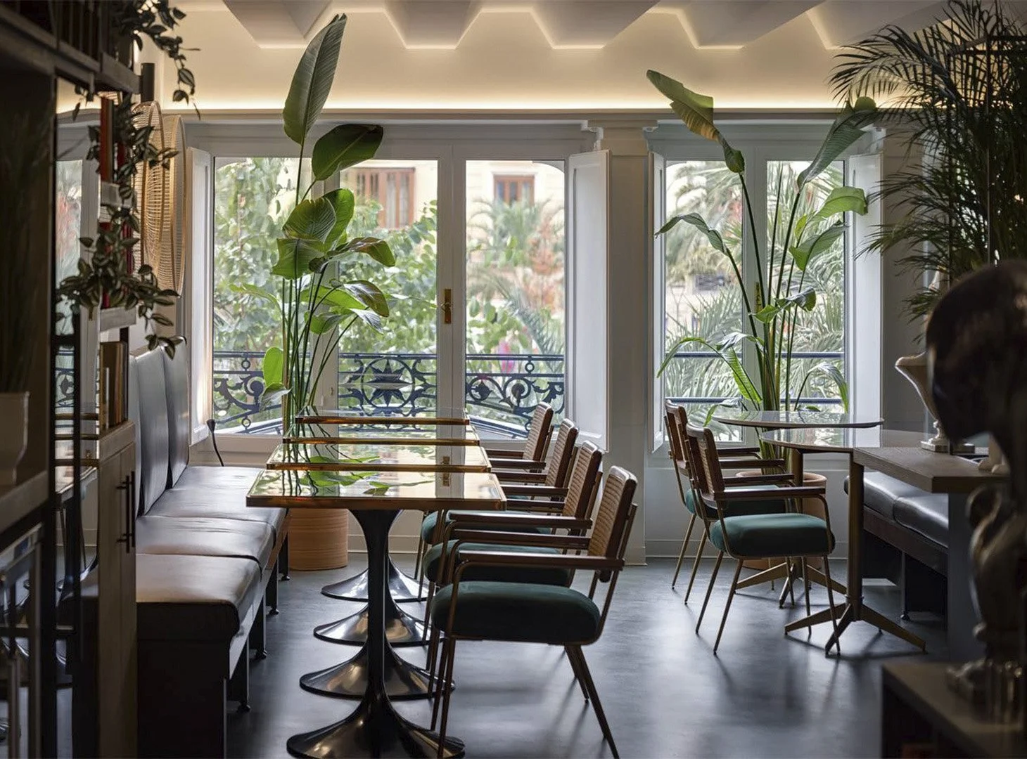 Interior of a bright, modern cafe with large windows and lush green plants, wooden chairs, and tables.
