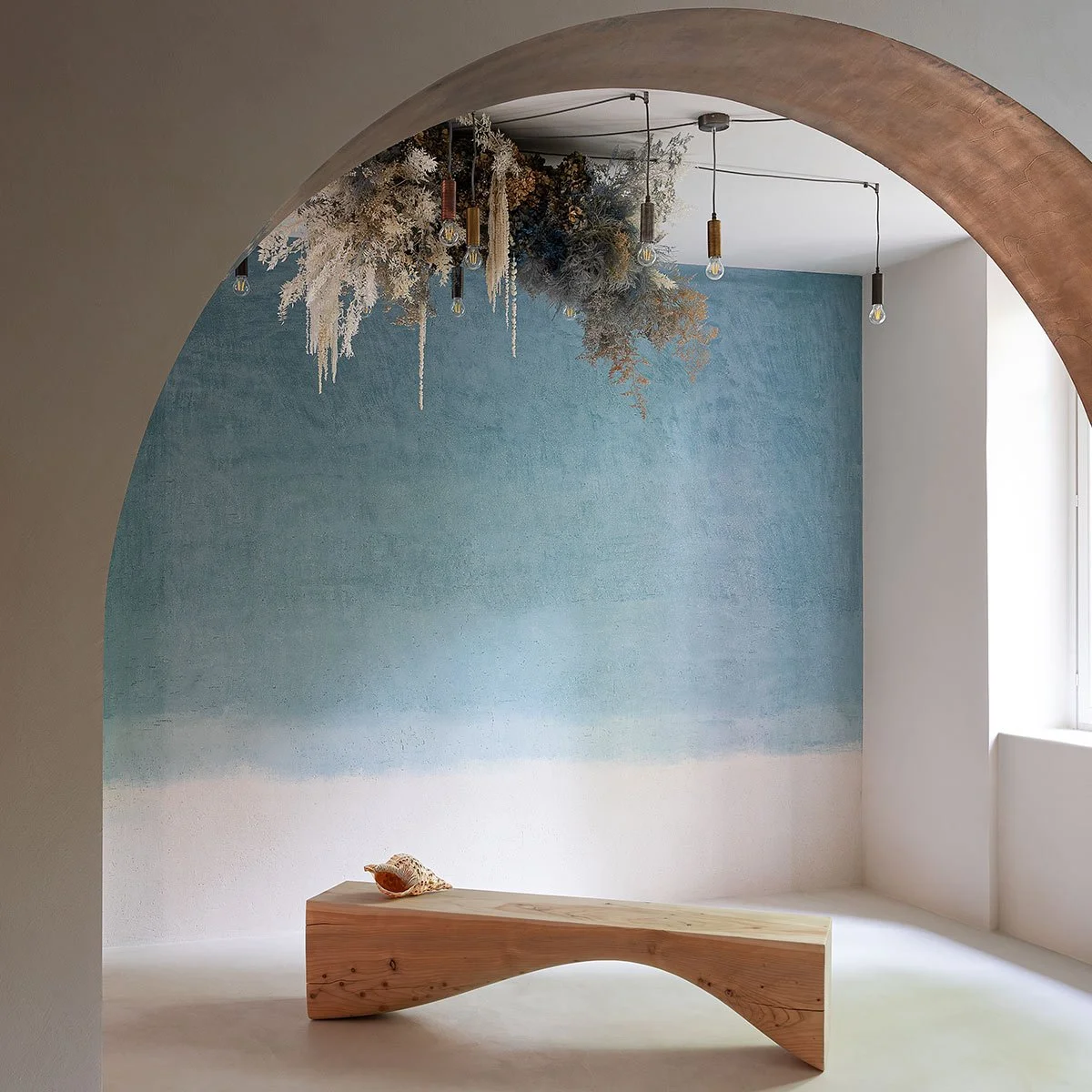 Minimalist room with a blue gradient wall, a small wooden bench with a seashell, hanging dried flowers and plants from the ceiling, pendant lights, and a large window providing natural light.