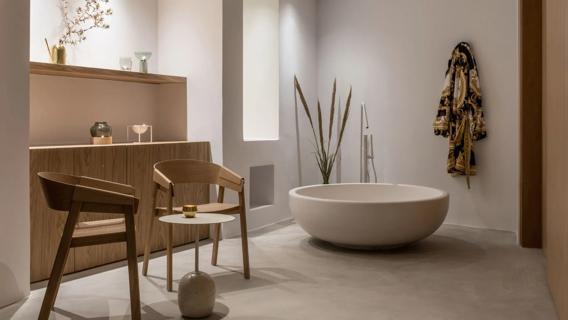 A minimalist bathroom with a large oval bathtub, light wood furniture, a small round side table with a candle, dried plants, a robe hanging on the wall, and decorative vases and candles on shelves.