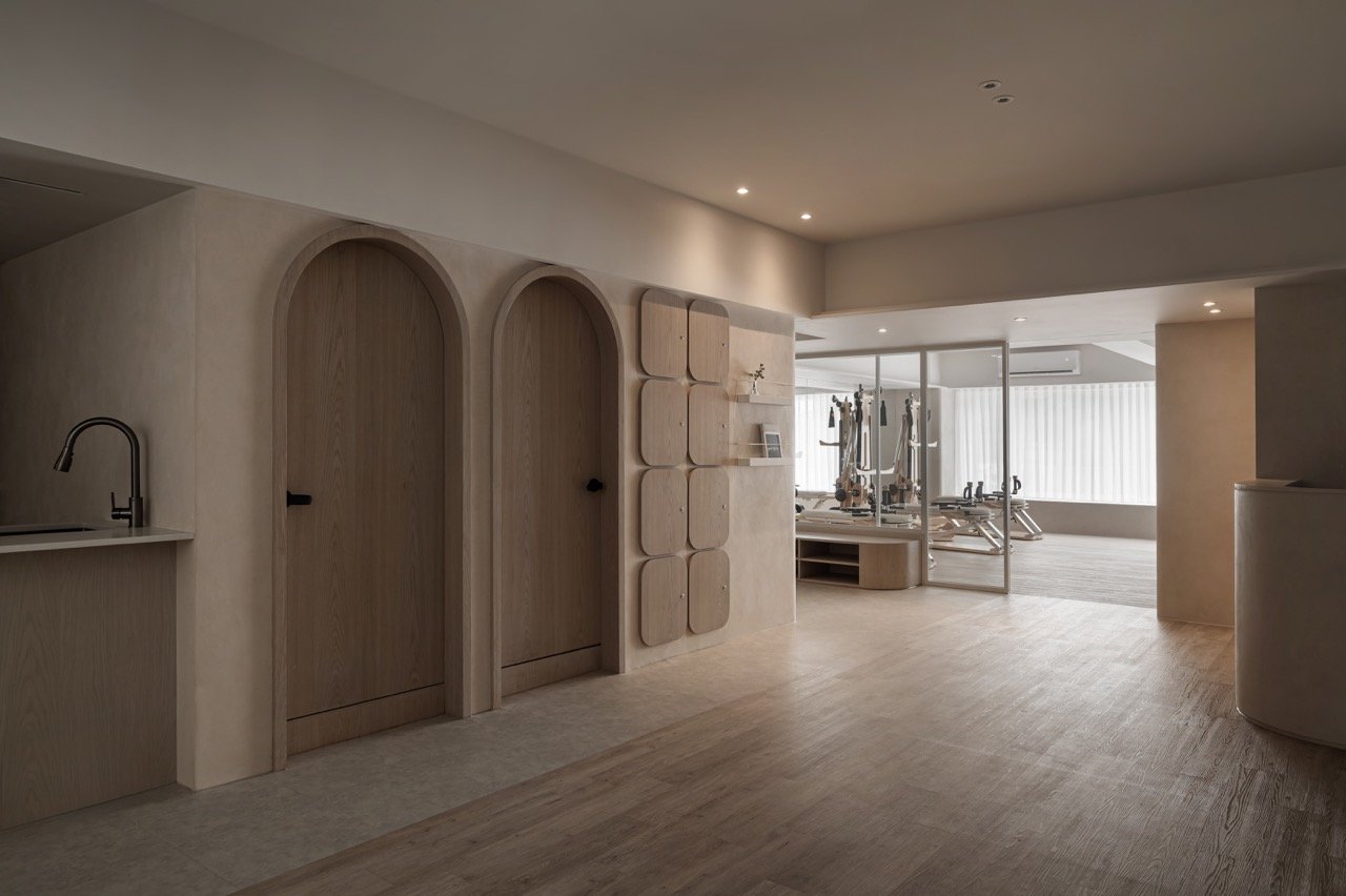 A minimalist microcement living space with light wood finishes, arched doorways, and a view into a gym area with workout equipment, separated by glass partitions.