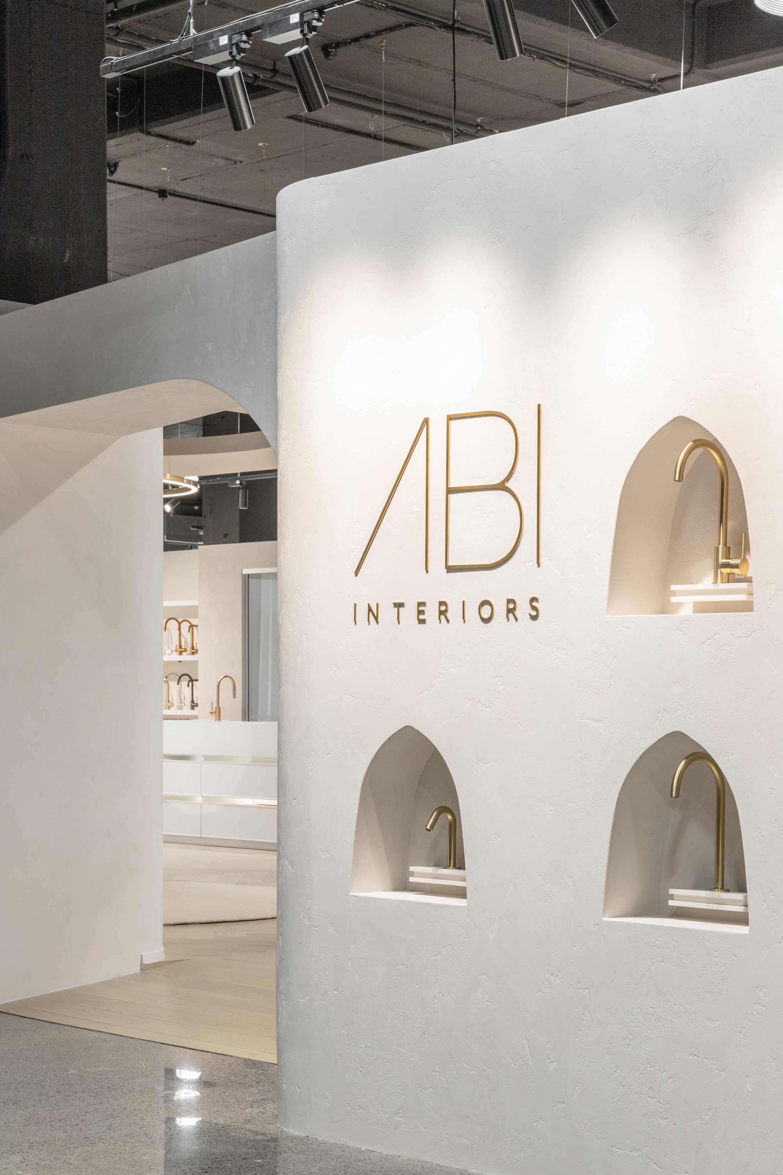 Interior of a showroom for ABI Interiors, featuring a white wall with gold text and decorative arches with gold faucets.