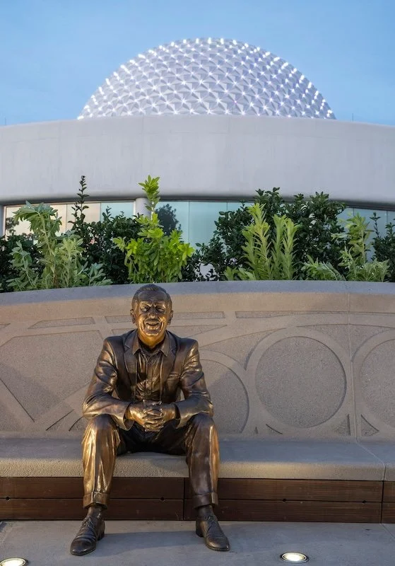 Bronze statue of a man sitting on a bench, smiling, against a backdrop of green plants and a modern building with a geodesic dome.