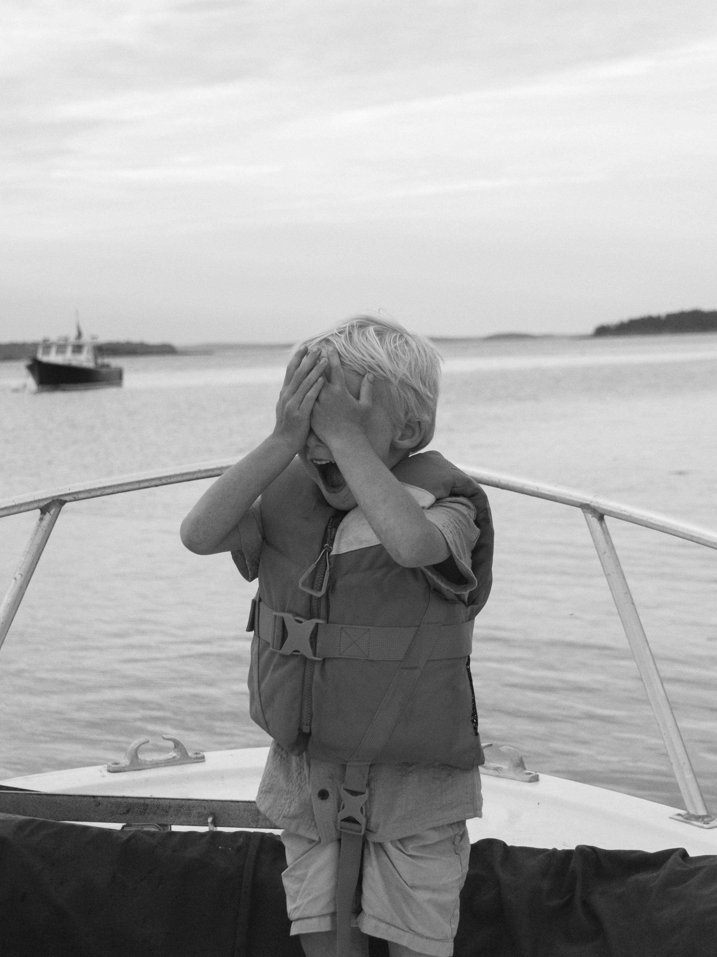 A young boy with blonde hair covering his eyes with his hands, standing on a boat with water and a distant boat in the background, appearing to be laughing or crying.