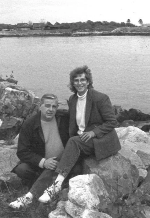 A black-and-white photo of a man and woman sitting on rocks near a body of water, with houses and trees in the background. The woman is sitting on the rocks, smiling, and the man is sitting beside her, holding her foot.