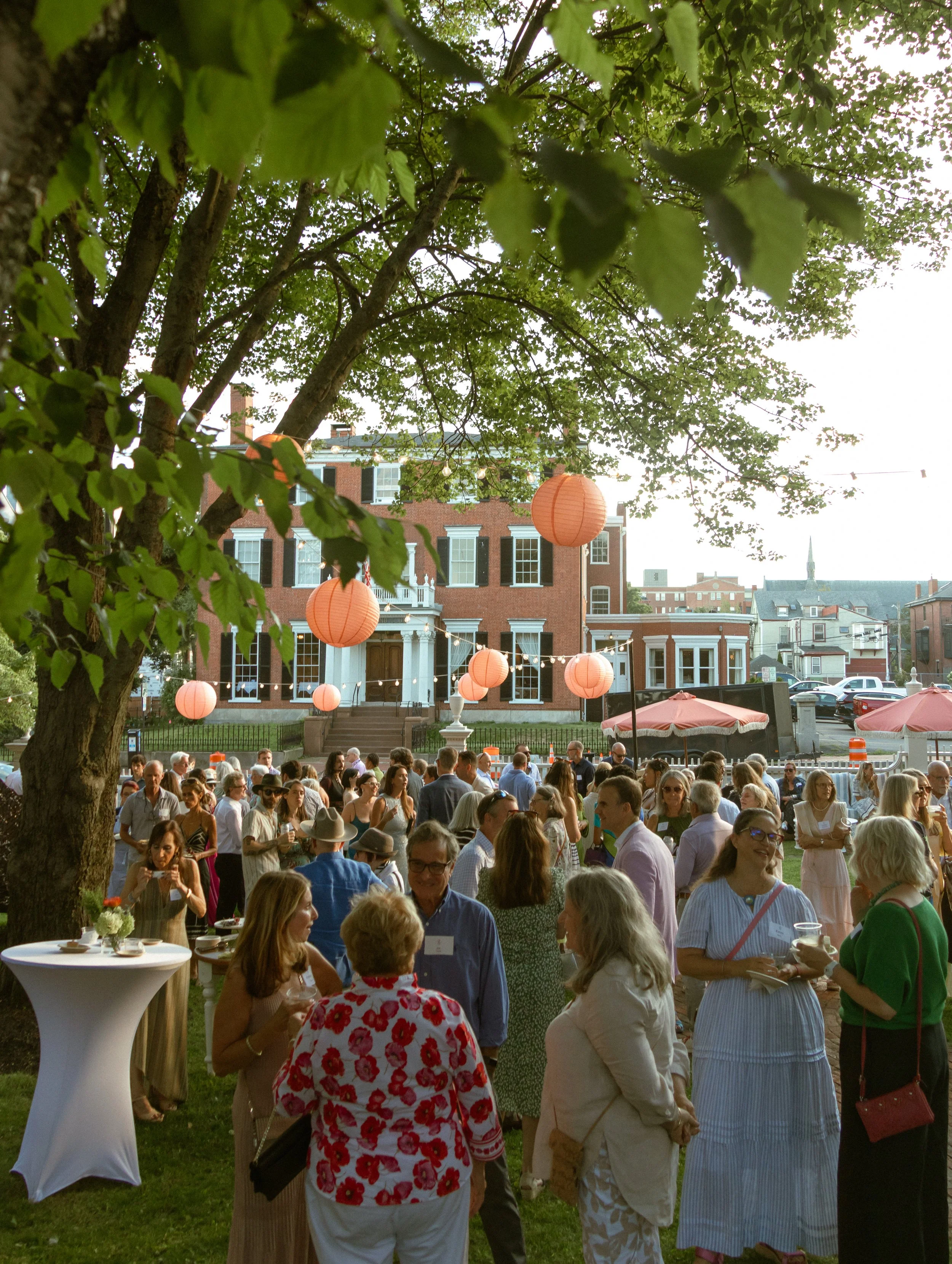 Crowd of people gathered outdoors at an event in front of a historic brick building with large windows, decorated with pink paper lanterns hanging from a tree, on a sunny day.