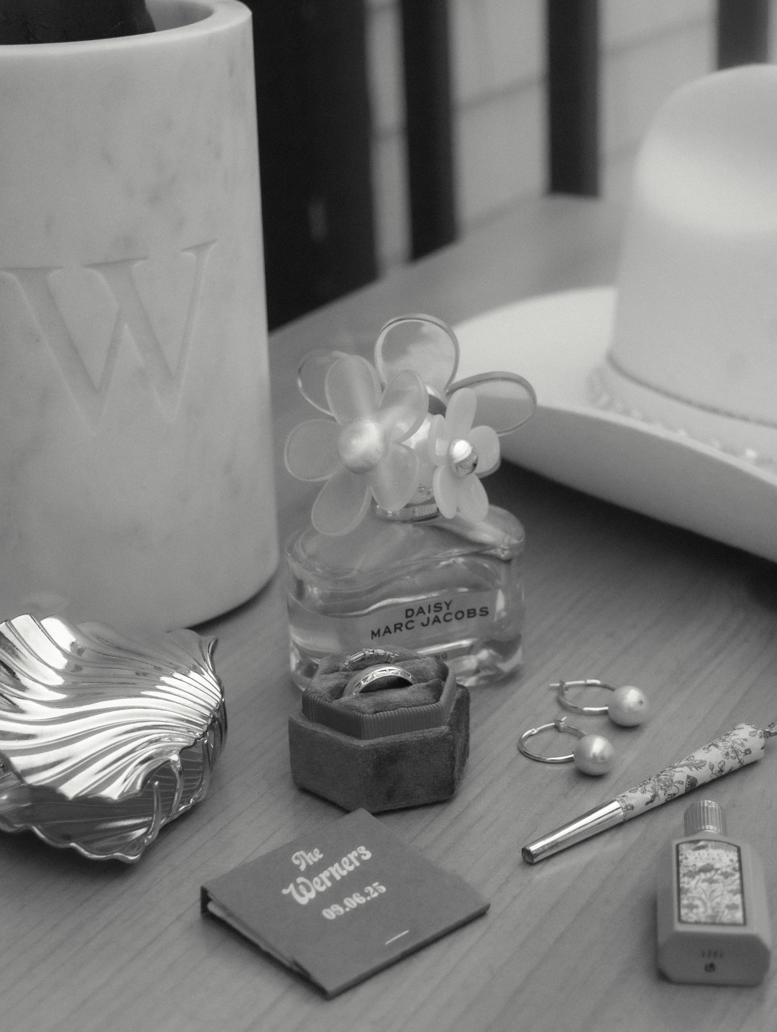 Assorted items on a table, including a large mug with letter W, a small perfume bottle with a flower-shaped cap labeled 'Daisy Marc Jacobs,' a ring in a small box, a pair of pearl earrings, a decorative pen, a small spray bottle, a chocolate box labe