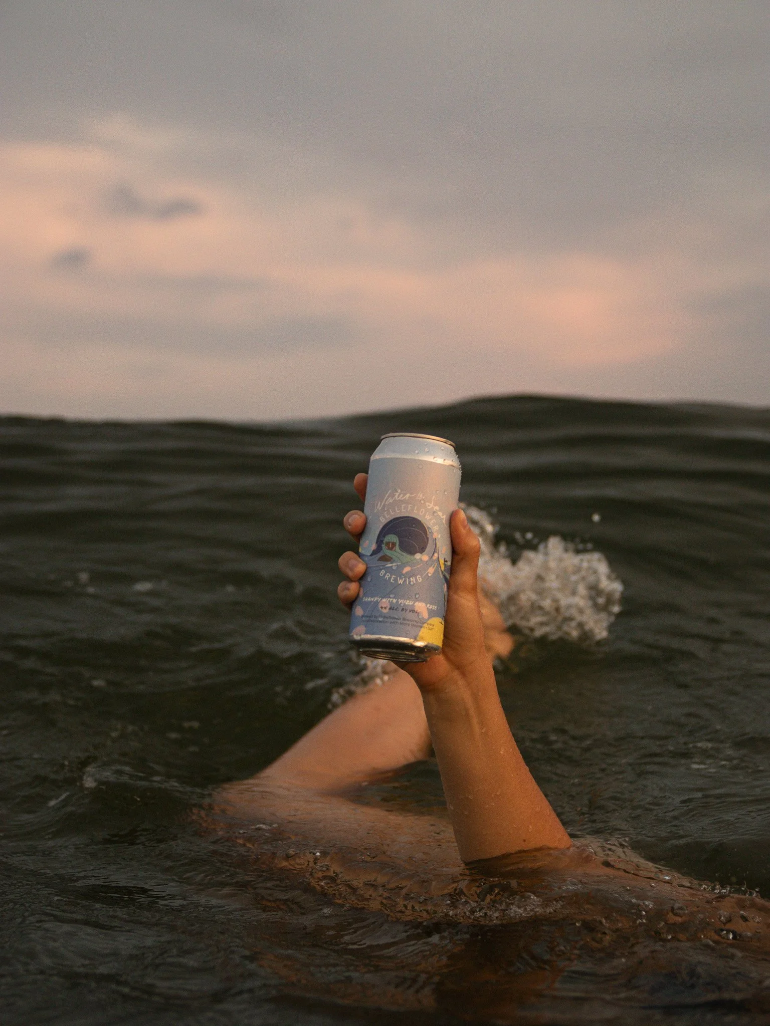 Person floating in water holding a can of beverage with an illustrated label, during sunset or dusk.