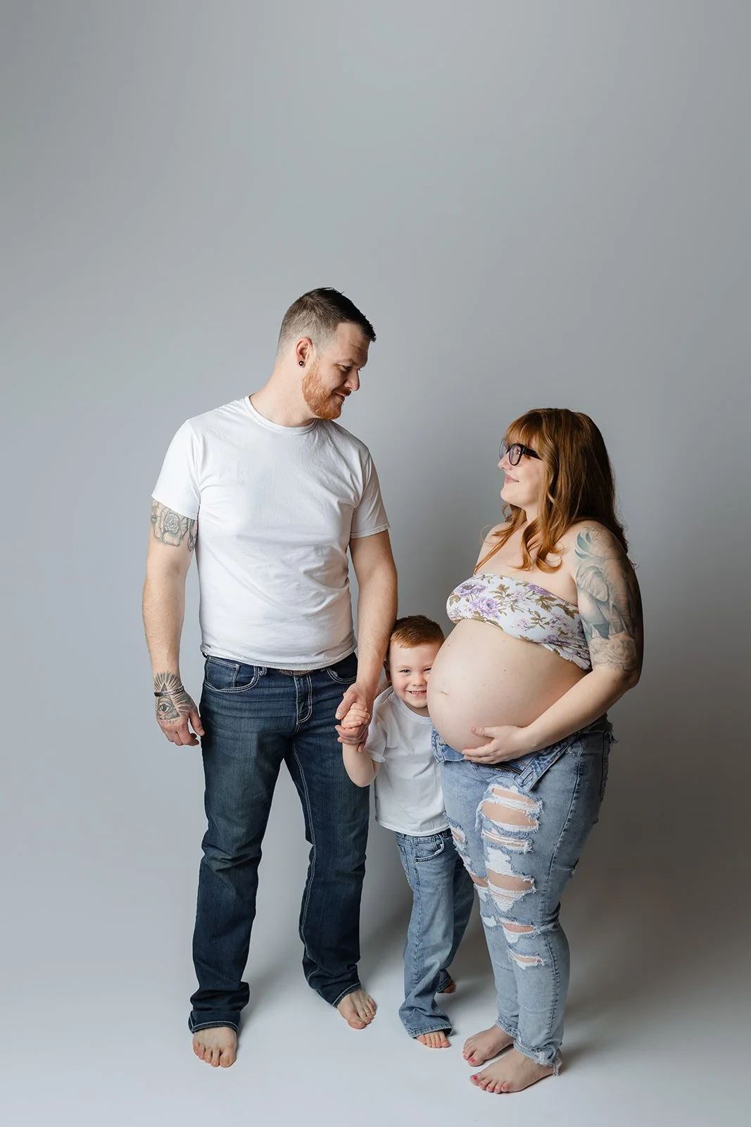 A family of three, including a pregnant woman, a man, and a young boy, standing together against a plain gray backdrop, holding hands and smiling.