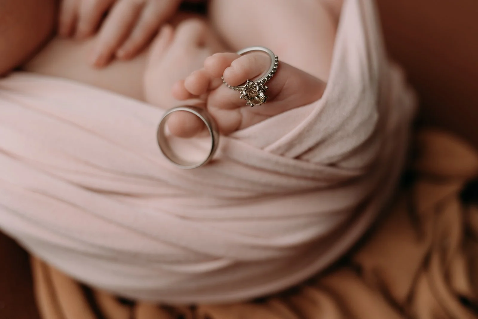Close-up of a hand holding two rings, one with a large central gemstone and smaller surrounding stones, and the other a plain band, against a soft pink fabric.