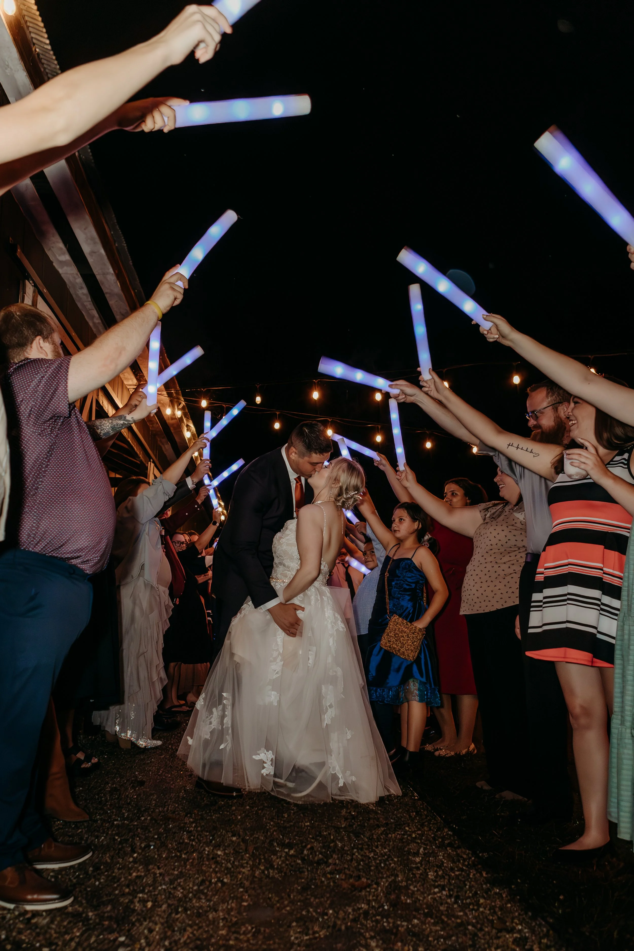 Bride and groom kissing under a canopy of light sticks held by guests at night during a wedding celebration.