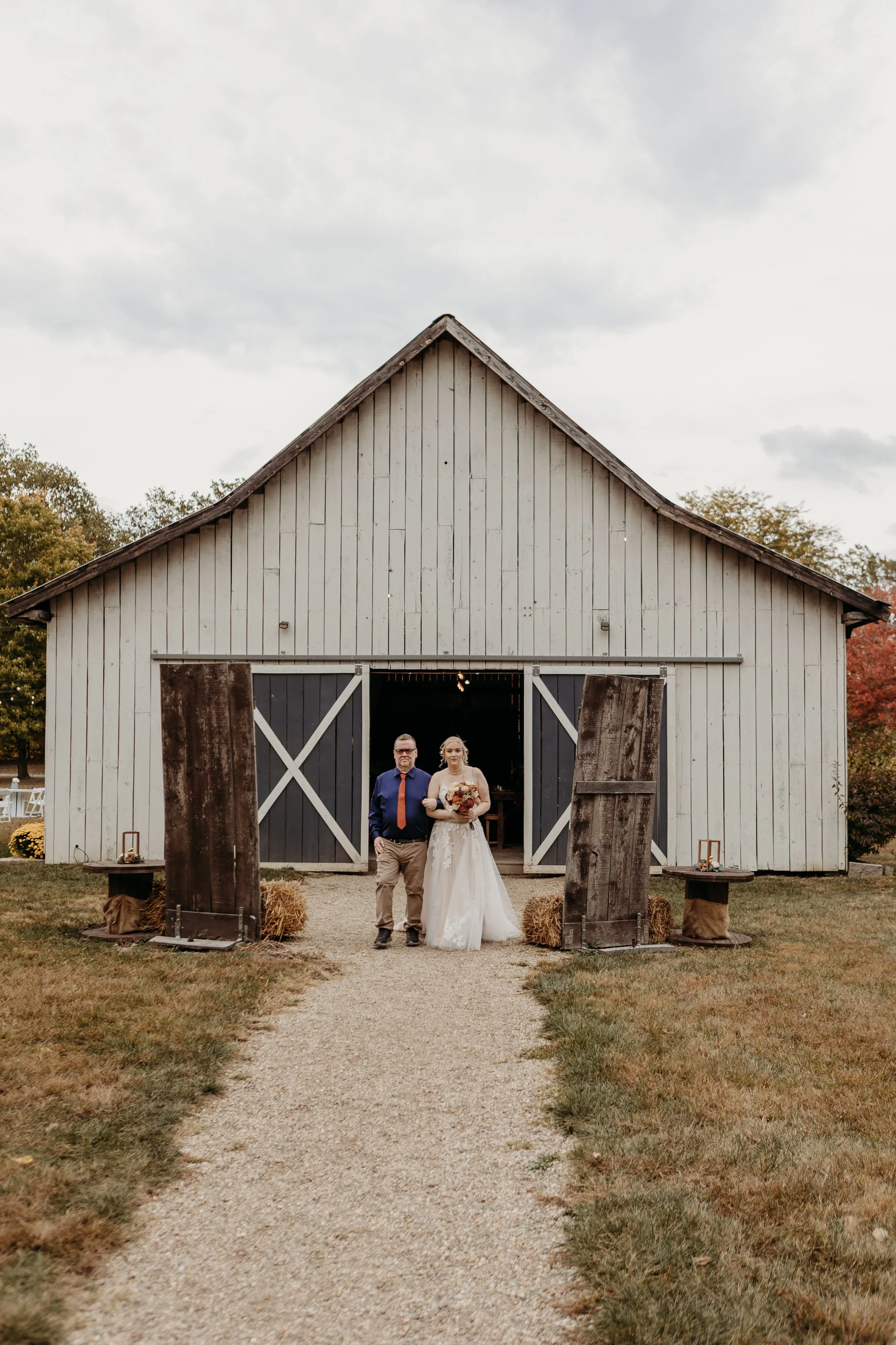 A bride in a white wedding dress walking with a man in a blue shirt and red tie out of a rustic barn, holding a bouquet of flowers, on a gravel path with hay bales and decorative tables on either side, under a cloudy sky.