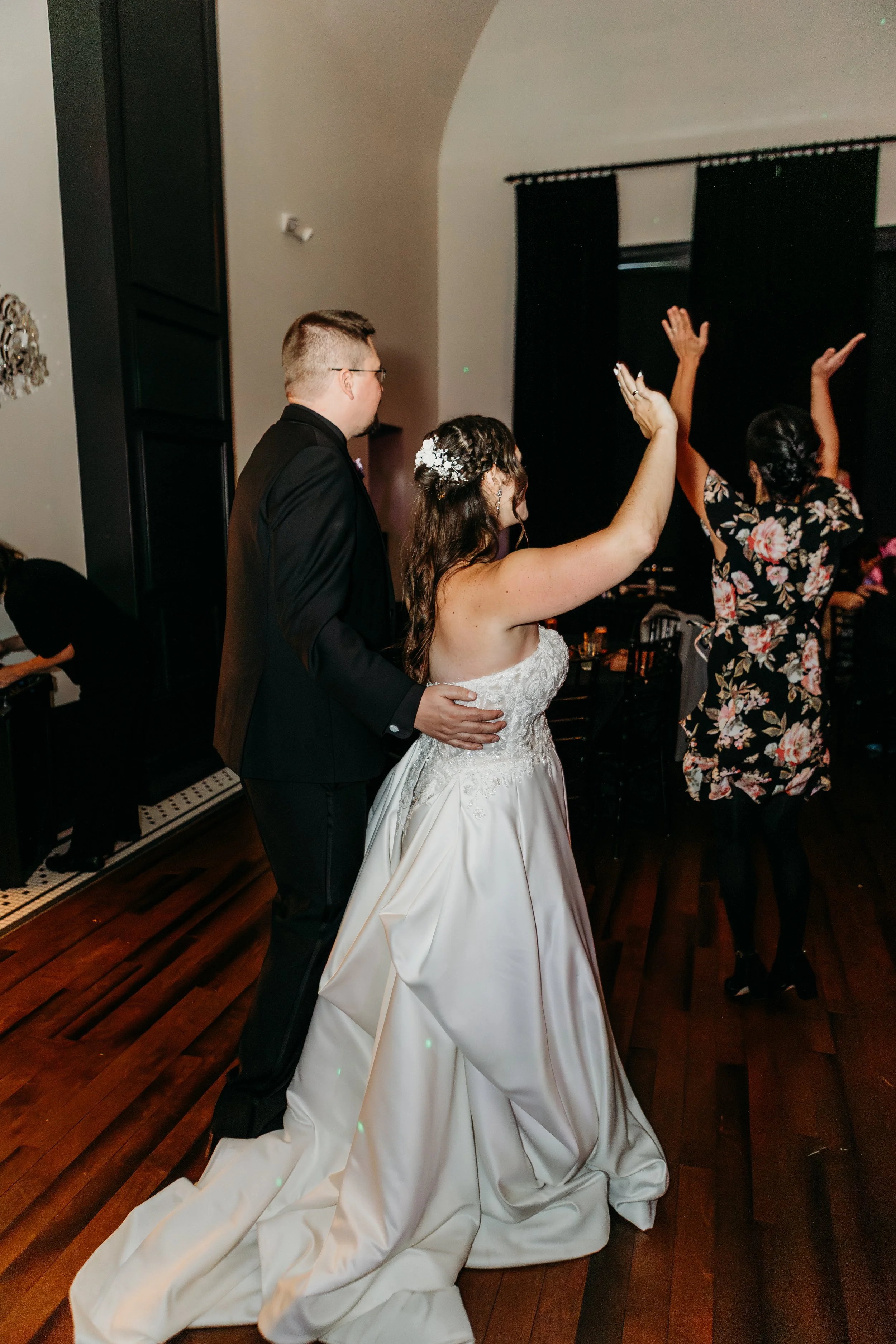 Bride and groom dancing at wedding reception, bride in white gown with floral hairpiece, groom in black suit, woman in floral dress dancing in the background.