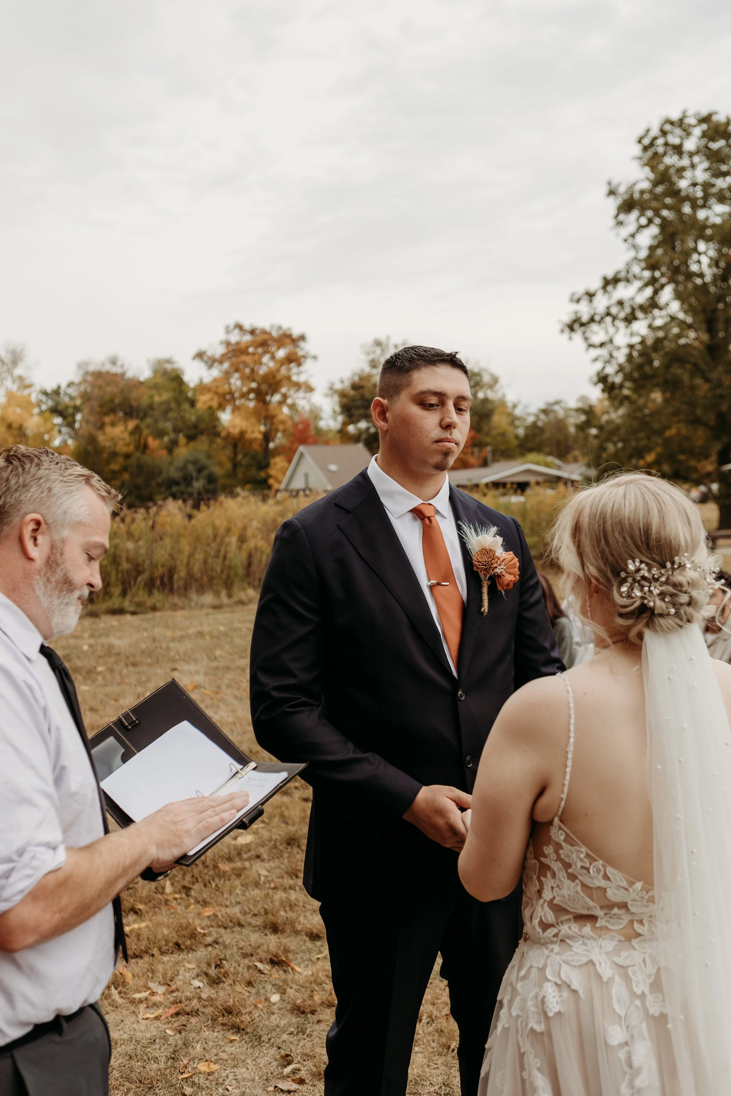 A couple getting married outdoors, facing each other, with a officiant taking notes nearby. The groom is wearing a suit with a boutonniere, and the bride is in a lace wedding dress with a floral hairpiece and veil. In the background, there are trees 