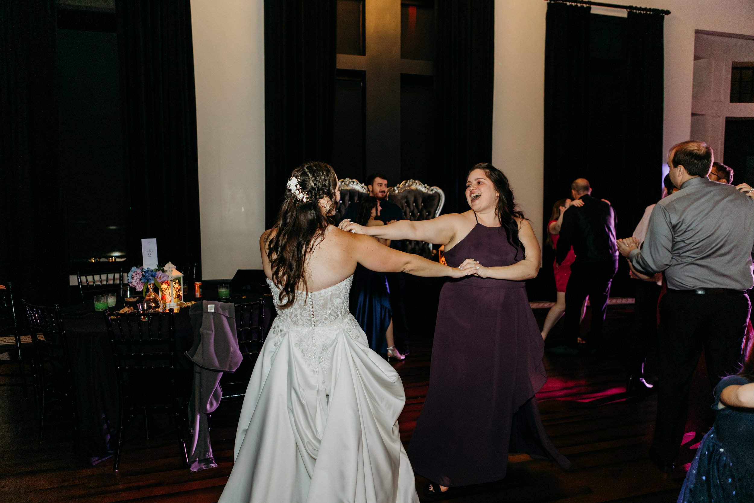 A bride and a woman dancing at a wedding reception, with other couples dancing in the background.