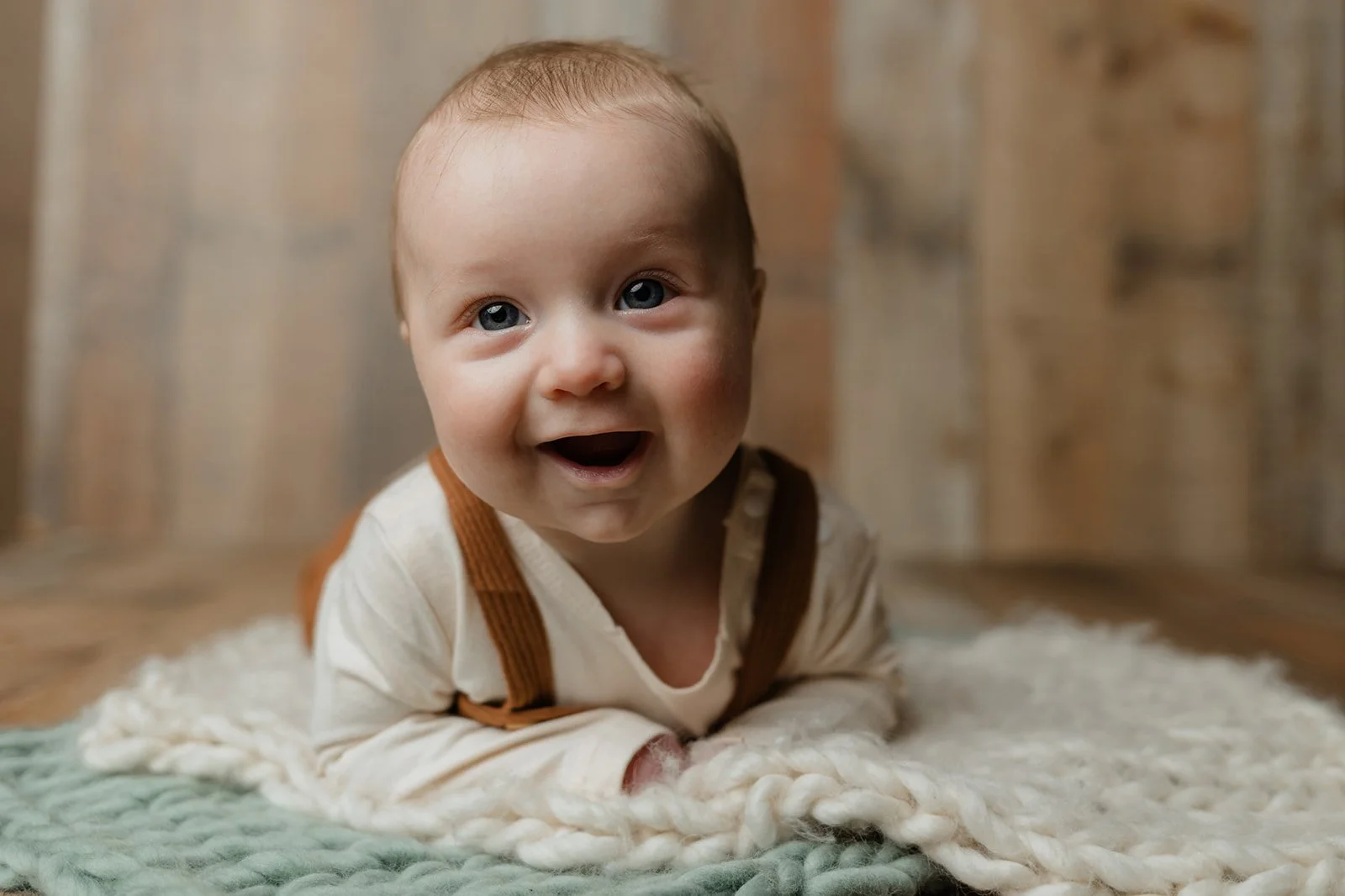 A smiling baby with blue eyes and light brown hair lying on a cozy, cream-colored rug with a wooden background.
