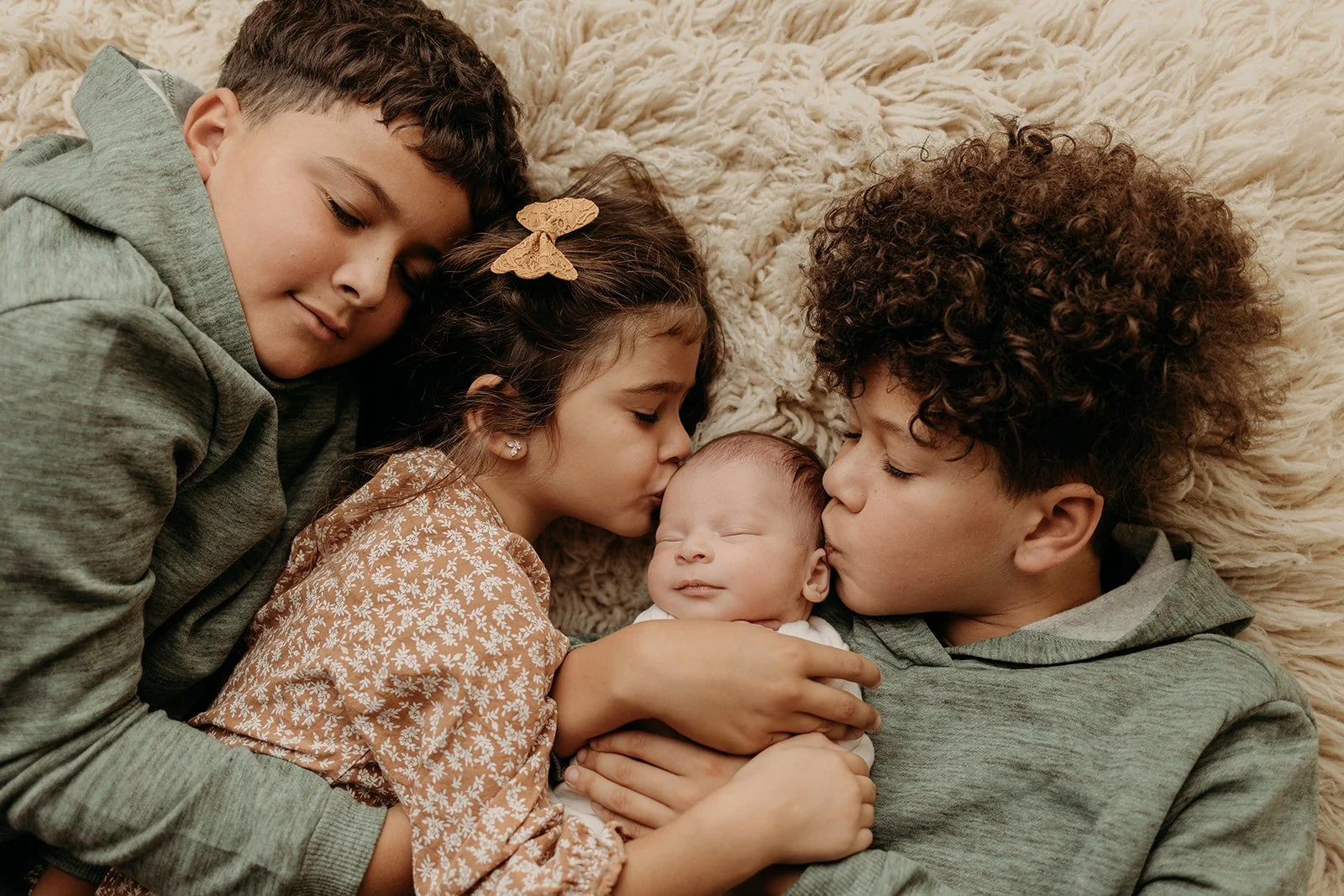 Four children lying on a fluffy beige blanket, with the youngest baby at the center being kissed on the head and cheek by her older siblings.