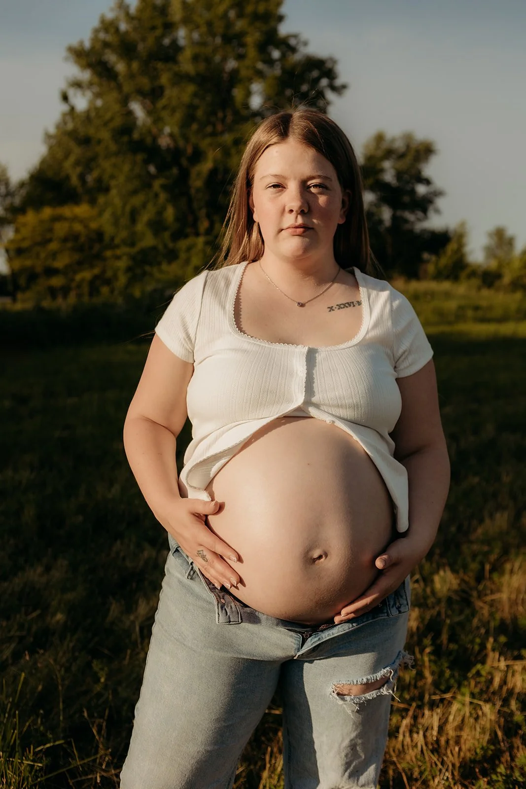 A young woman with long brown hair standing outdoors at sunset, holding her pregnant belly with both hands, wearing a white crop top and ripped jeans.