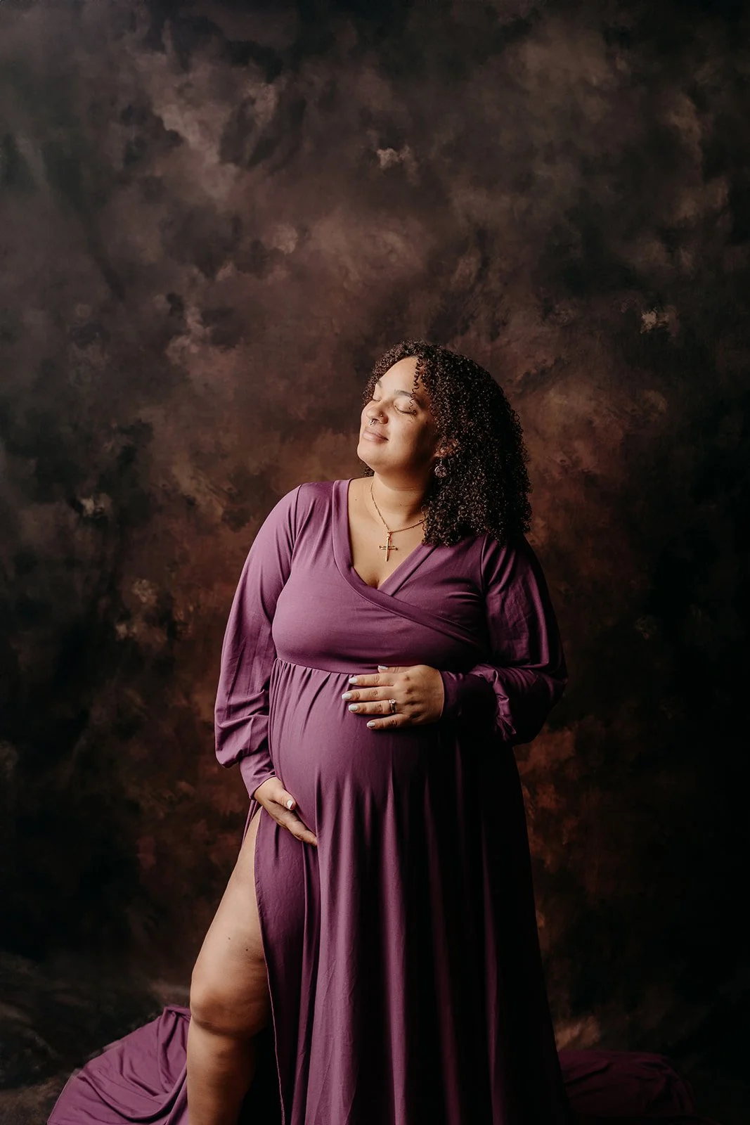 Pregnant woman standing with eyes closed, wearing a long purple dress with a slit, and jewelry including a cross necklace, in front of a dark, textured background.