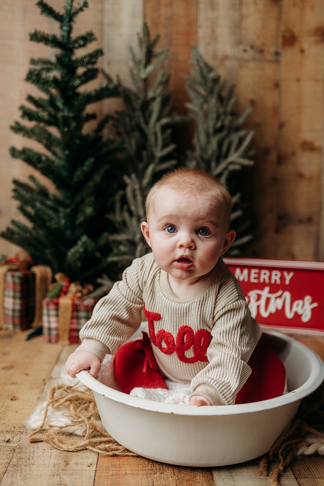 A baby in a cream sweater with red lettering, sitting in a white basin on a wooden floor with Christmas decor, including a green Christmas tree, wrapped presents, and a sign reading 'Merry Christmas' in the background.