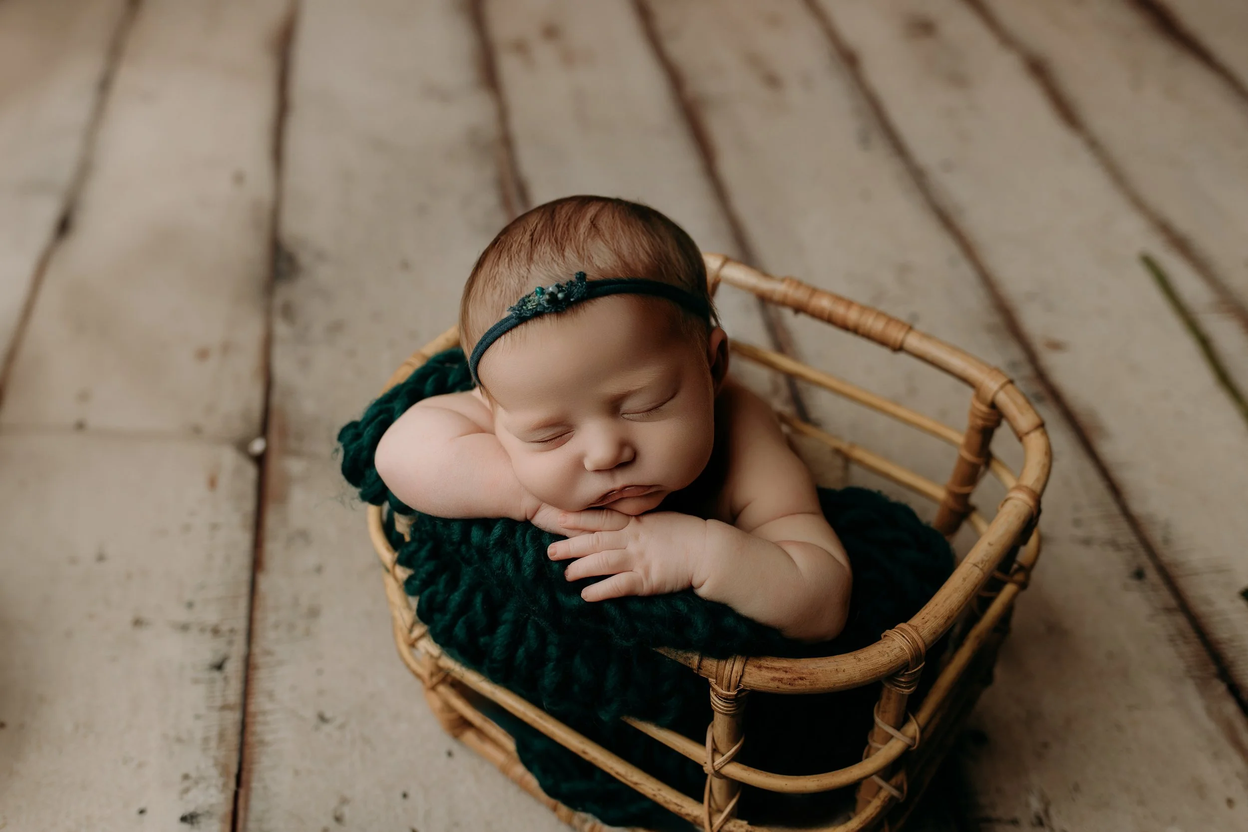 A sleeping newborn baby with a headband, lying on a green blanket inside a small wicker basket, on a wooden floor.