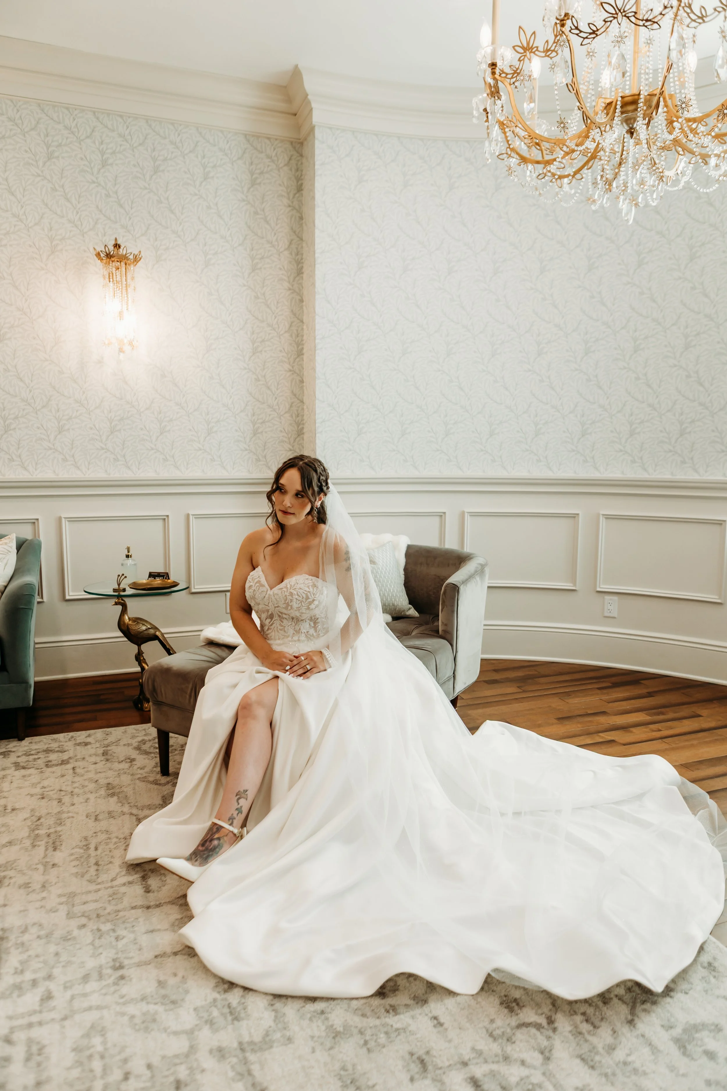 Bride in a wedding dress sitting on a gray sofa in a luxurious room with chandelier and ornate wall decor.