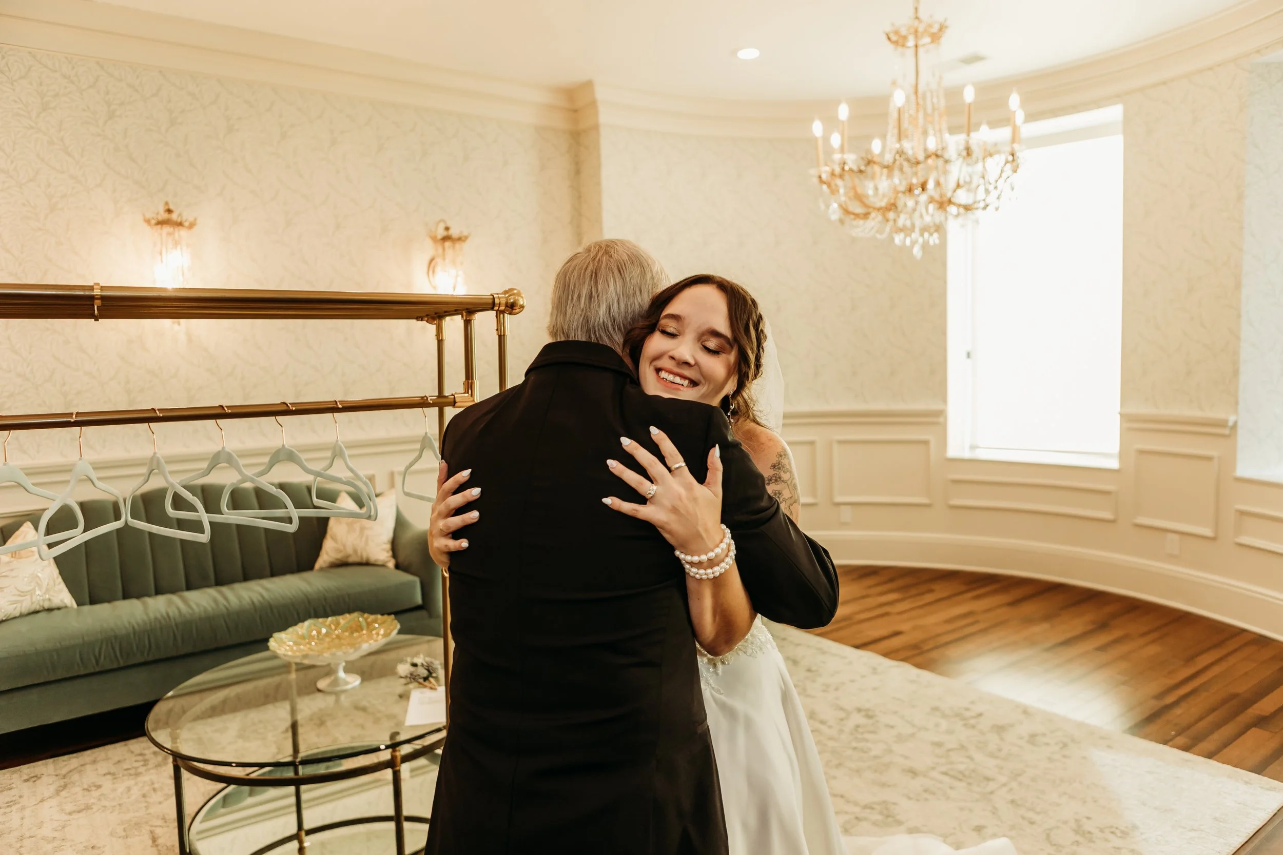 A bride hugging an older man in a room with elegant decor, a chandelier, and a green velvet sofa.
