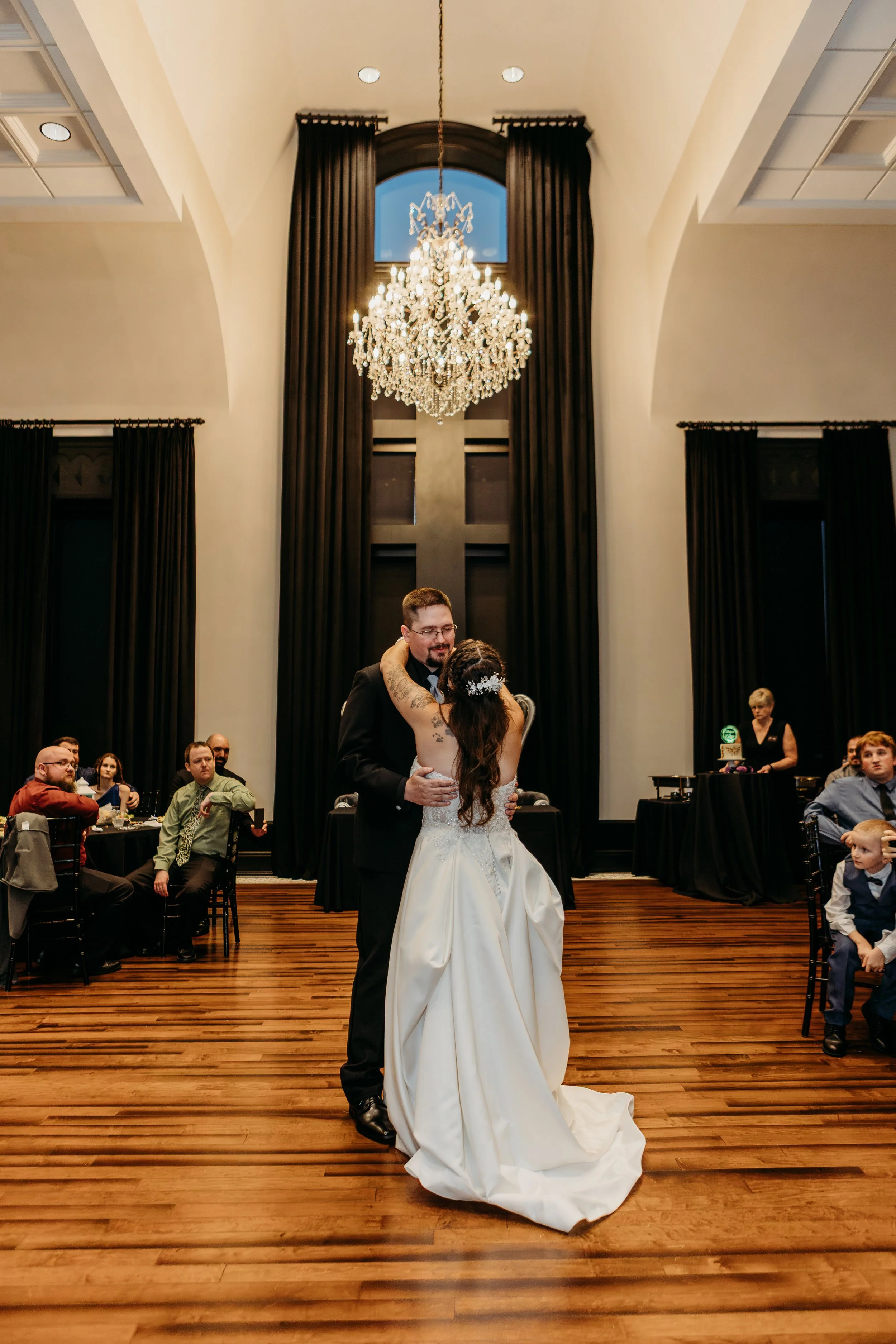 A bride and groom share their first dance at a wedding reception in a grand hall with a chandelier and tall black curtains.