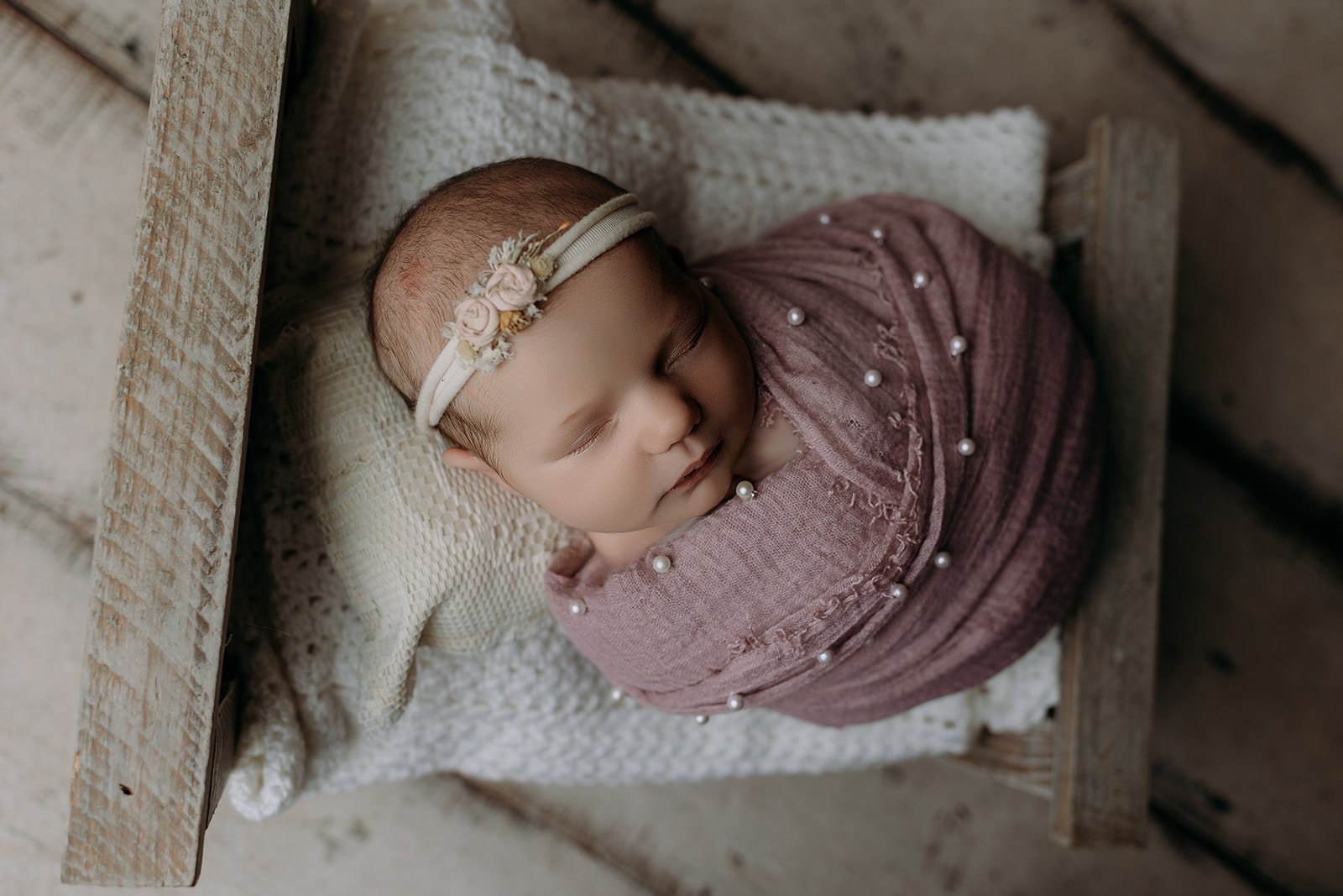 A sleeping baby girl with a floral headband, wrapped in pink fabric with pearls, lying in a small wooden bed with white knitted blanket and cream pillows.