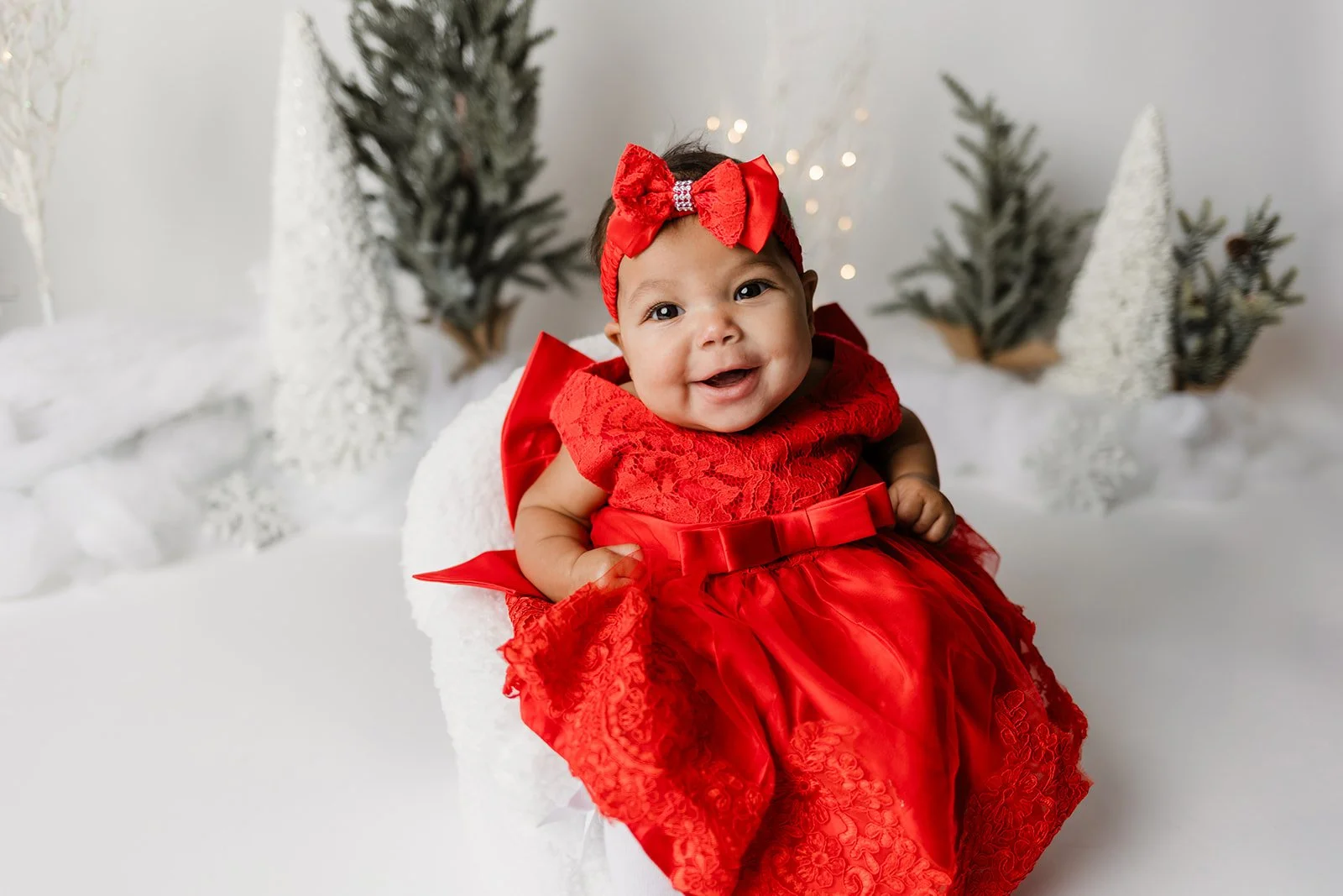 A smiling baby girl in a red lace dress with a satin bow, wearing a red headband with a bow, sitting in a white furry chair with a holiday-themed background of small decorated Christmas trees and snow.
