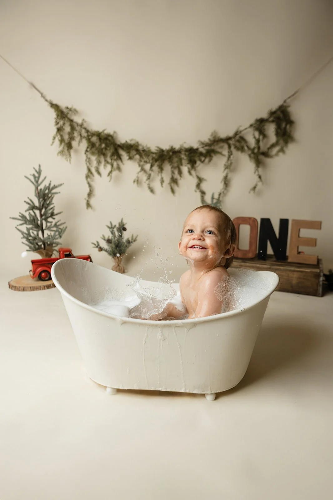 A smiling toddler taking a bath in a small vintage tub with a festive backdrop including two miniature Christmas trees, a wooden log with decorative letters spelling 'ONE,' and greenery hanging above.