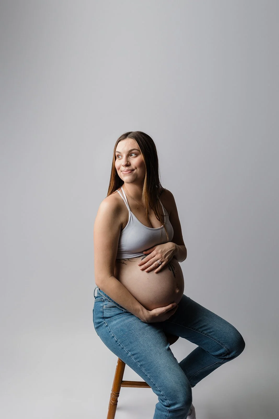 A pregnant woman sitting on a wooden stool against a plain grey background, wearing a white tank top and jeans, cradling her belly with both hands, smiling softly.