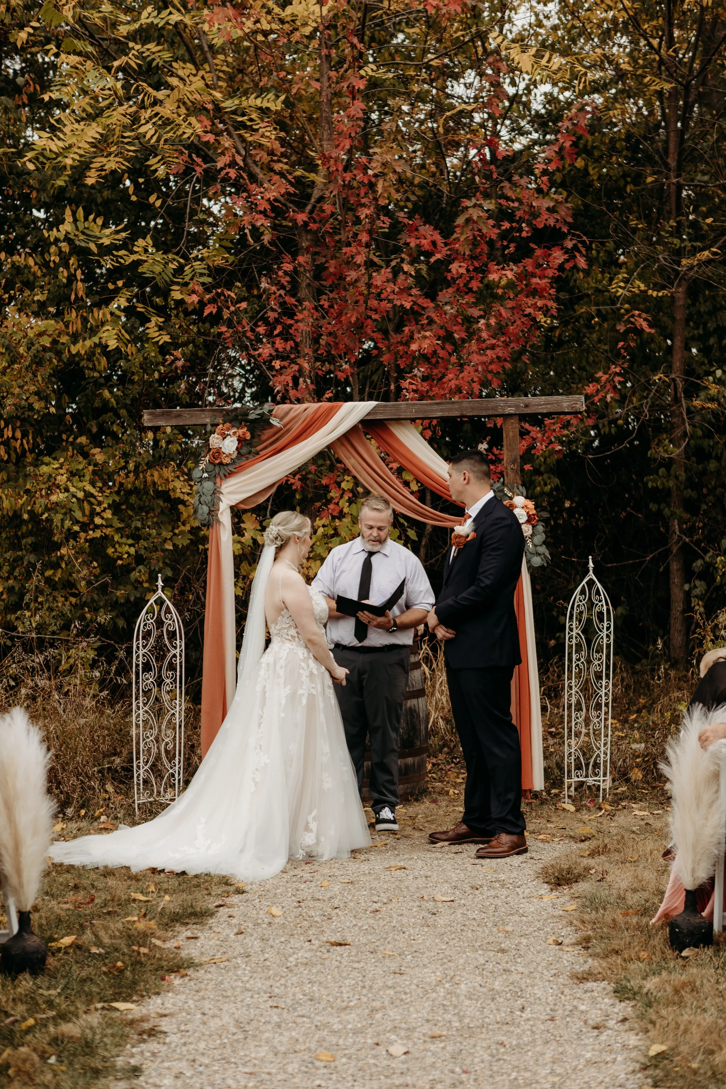 A couple gets married outdoors beneath a decorated wooden arch with fall-colored drapes and flowers, officiated by a man holding a book, surrounded by autumn trees and wedding guests.