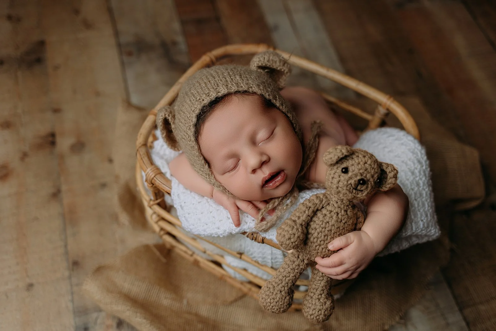 Adorable sleeping baby in a wicker basket, wearing a tan knit bear hat and holding a small tan knit teddy bear, on a wooden floor with burlap fabric underneath.
