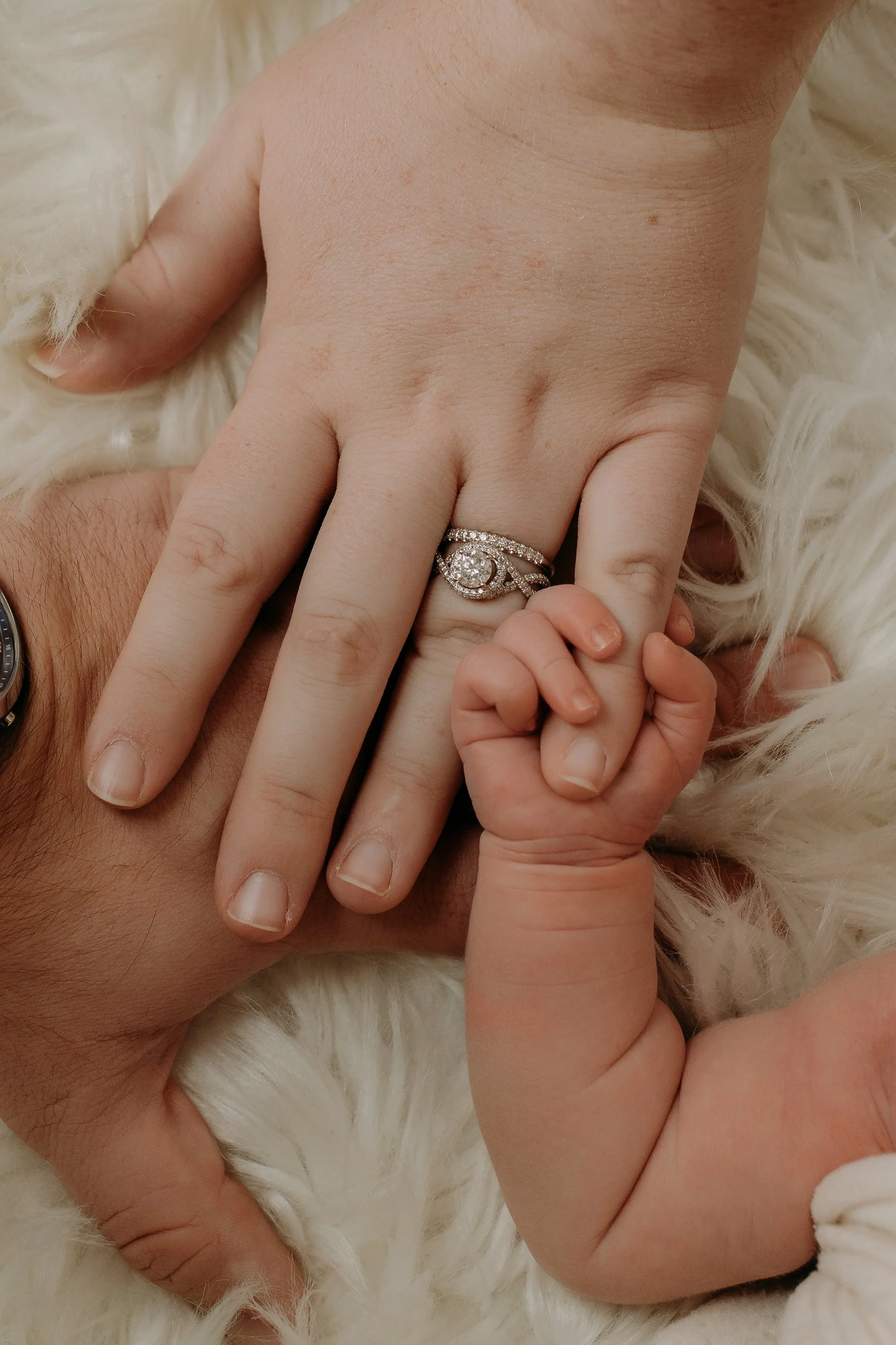 A close-up of a hand with wedding and engagement rings, holding a small child's hand on a fluffy white surface.