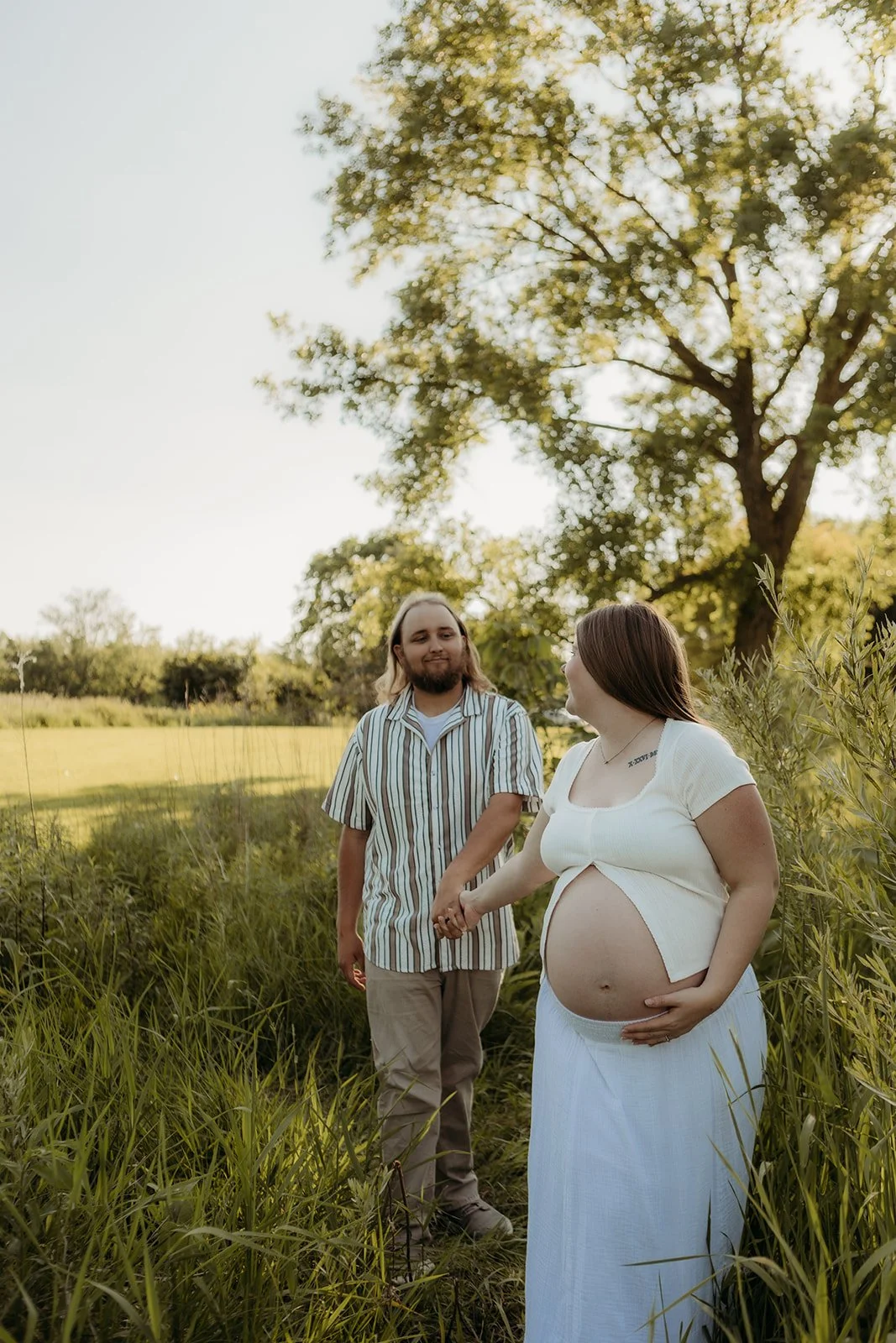 A pregnant woman holding her belly and a man holding her hand, standing in a grassy field with trees in the background during sunset.