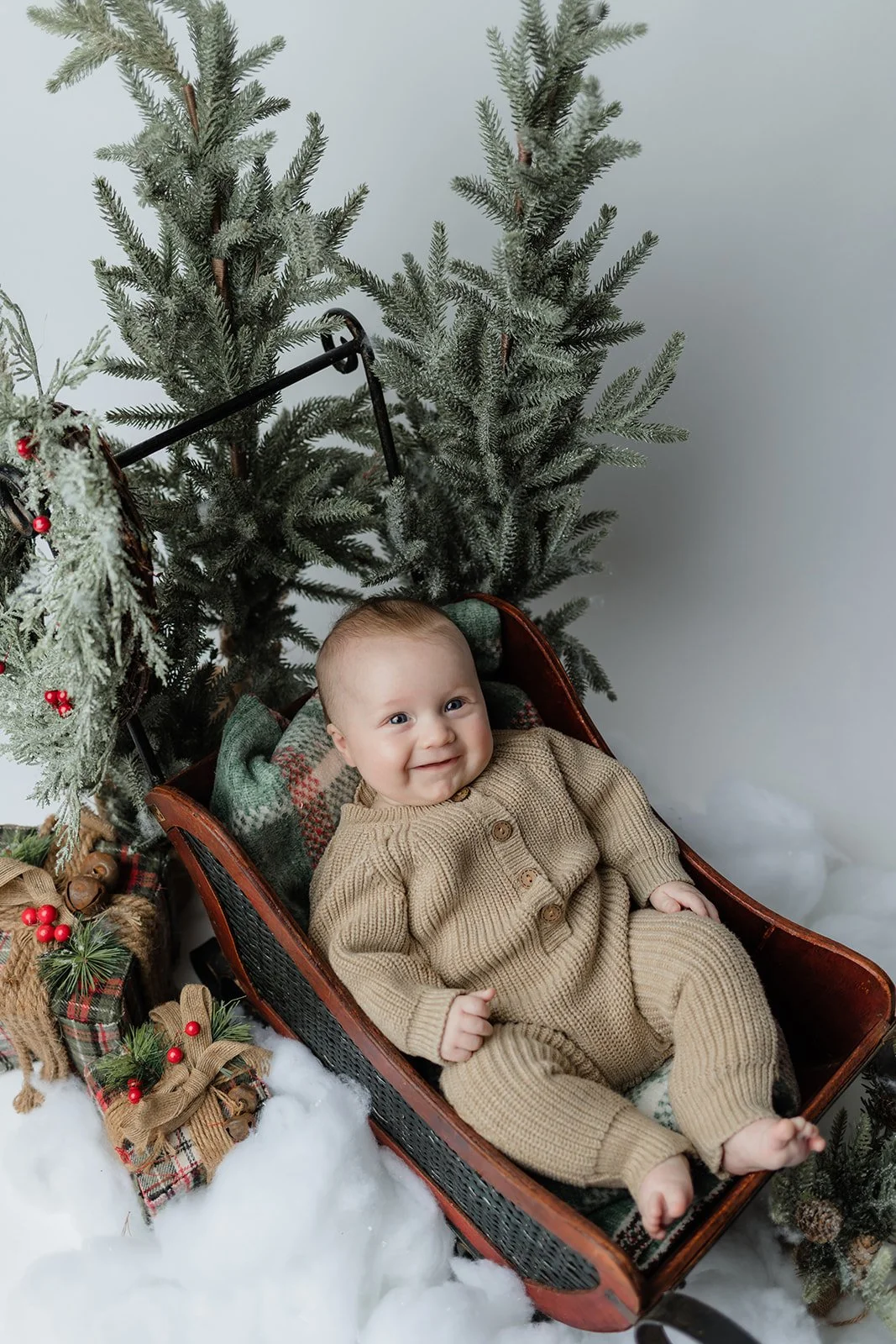 A baby lying in a sled surrounded by Christmas decorations and artificial snow, with a Christmas tree in the background.